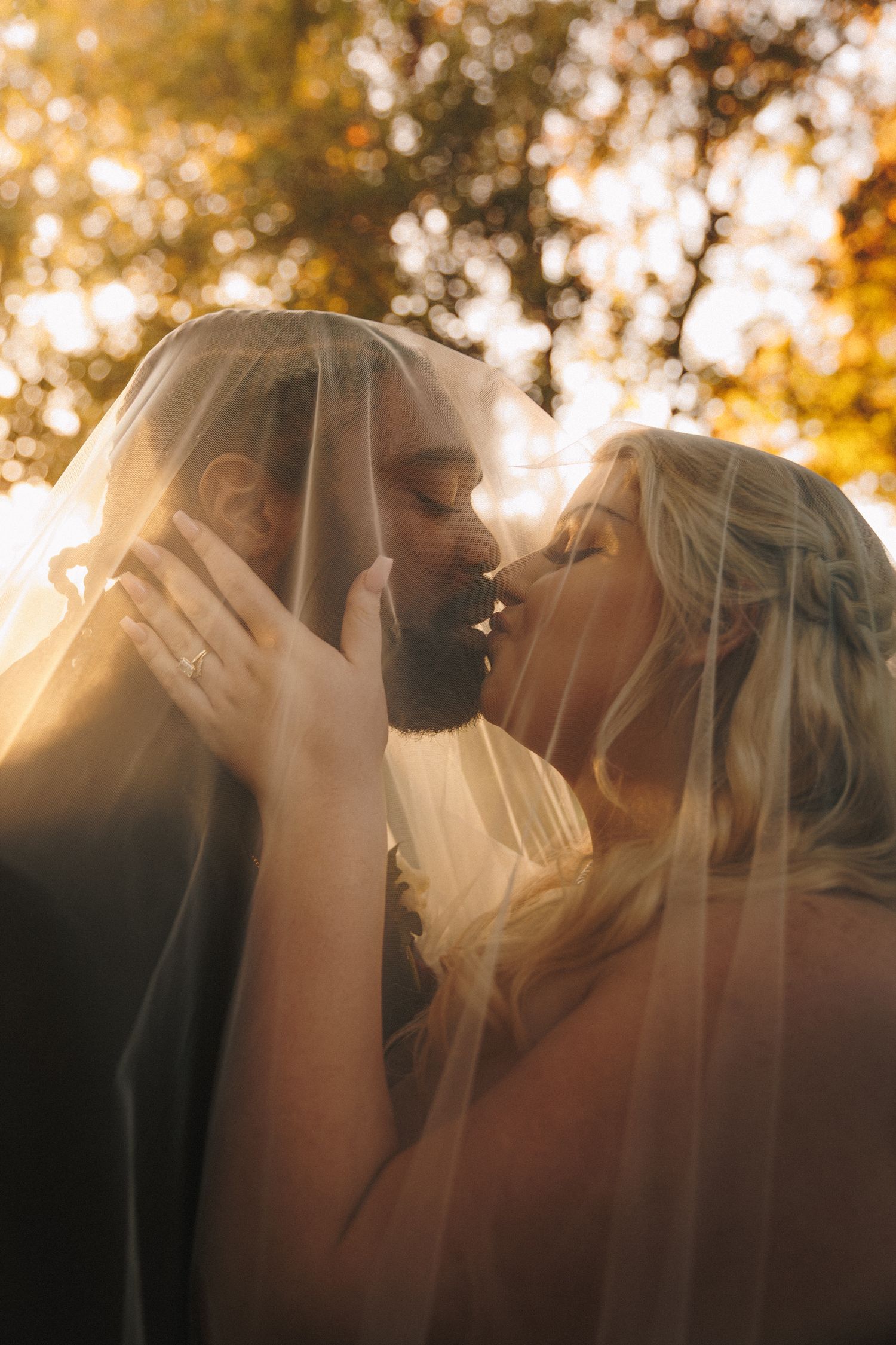 Bride and groom kissing under veil in golden autumn sunlight during wedding portraits.