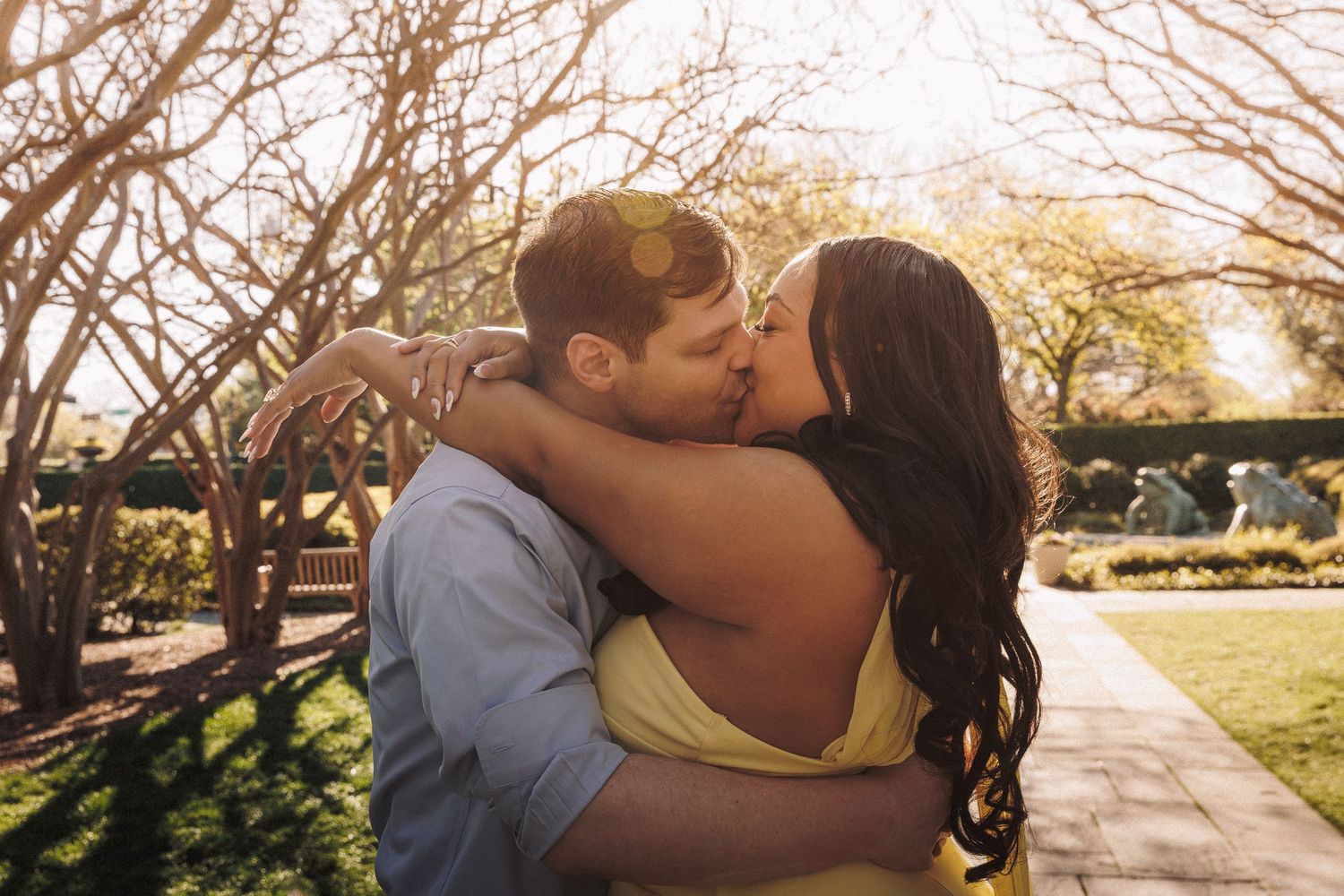 Couple sharing a romantic kiss in a sunny garden, surrounded by blooming trees and lush greenery.