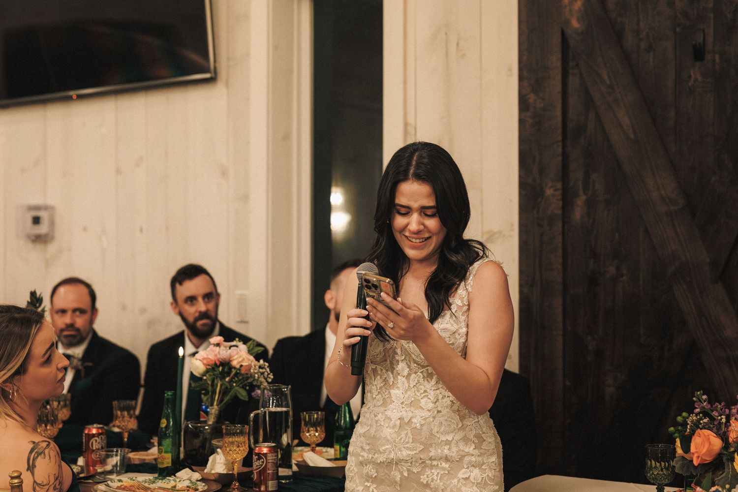 Bride in beaded gown smiles while reading from her phone during a speech at the wedding reception dinner.