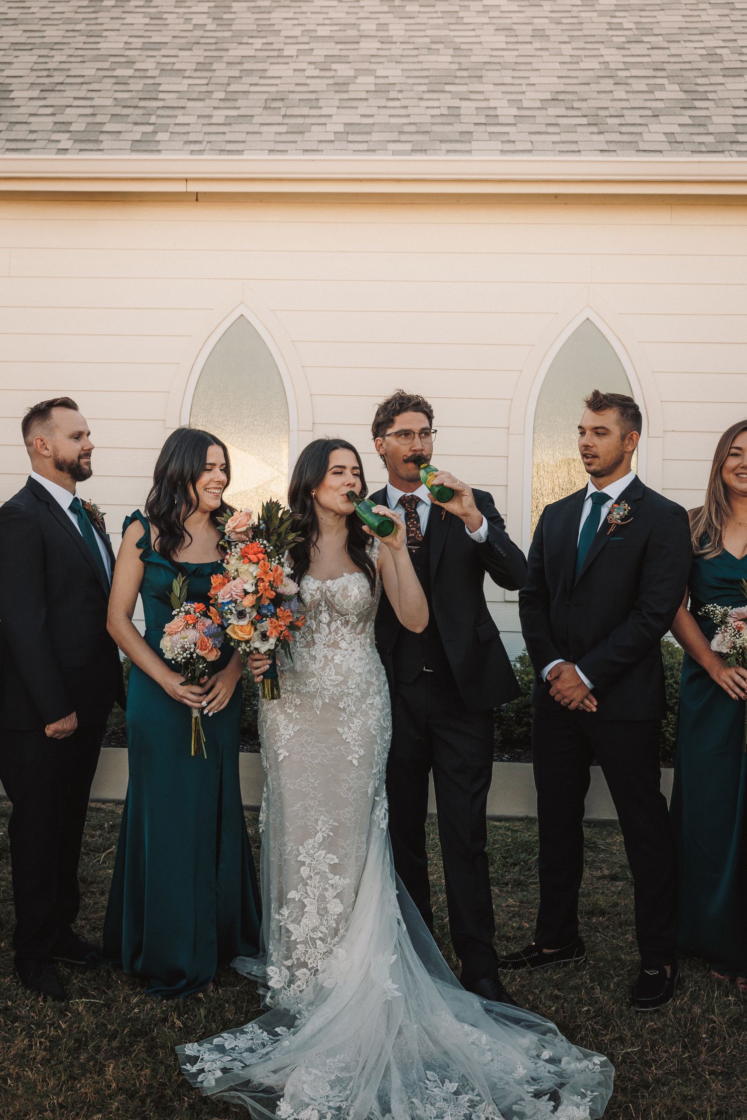 Wedding party in emerald and black attire celebrating outside a white chapel at sunset.