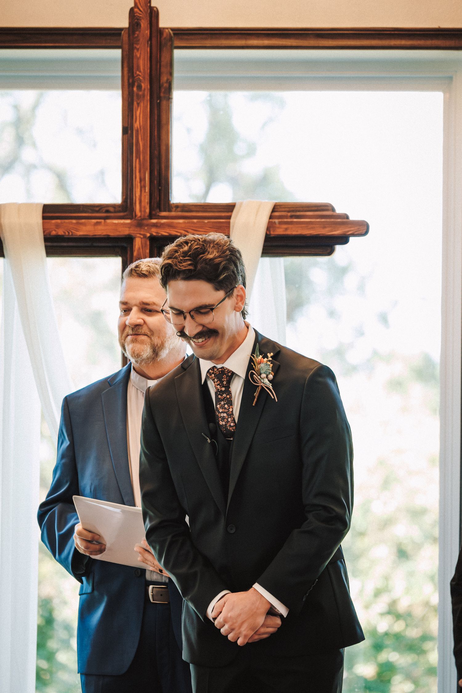 Groom with mustache smiles at altar beside officiant holding ceremony program, wooden cross backdrop.