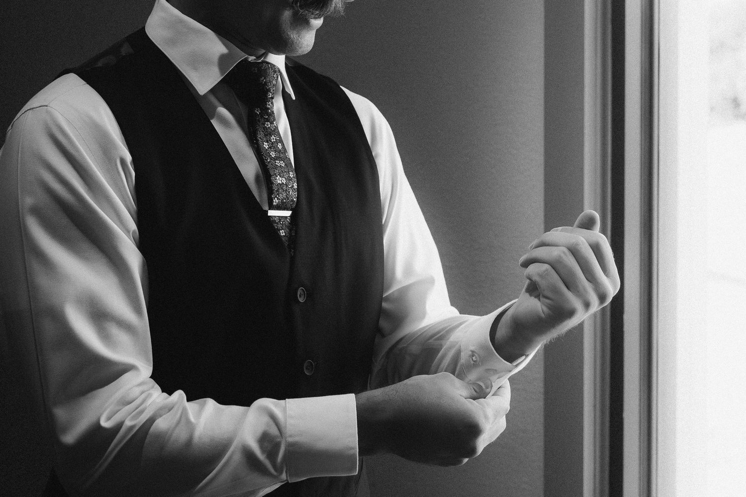 Groom in black vest and tie adjusting cufflinks by window in black and white photo.