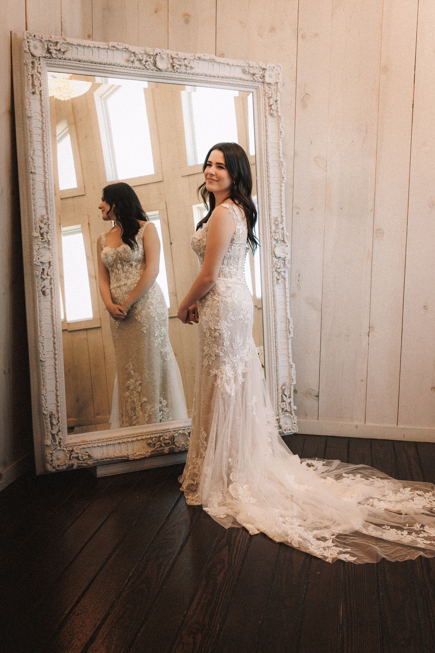 Bride in lace wedding gown with train poses beside ornate white mirror in elegant wood-paneled room.