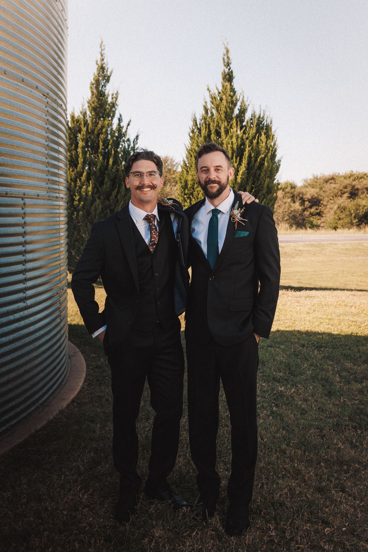 Two groomsmen in black suits smiling together outdoors near a grain silo and cypress trees on a sunny day.