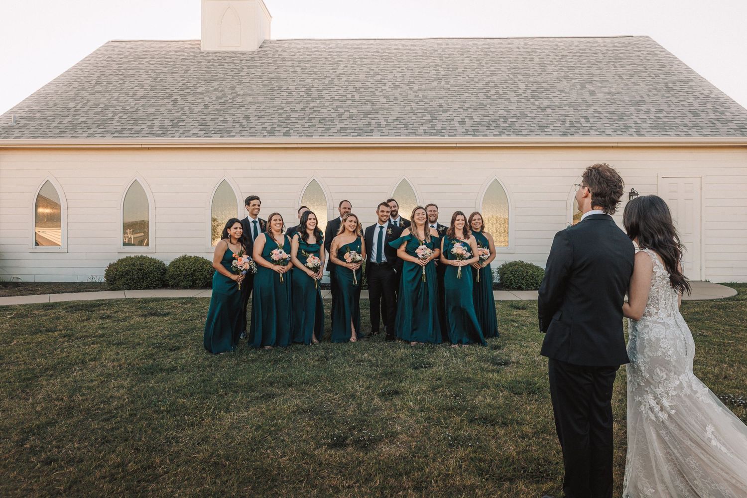 Wedding party in emerald green gowns poses outside a white chapel while bride and groom share a first look.