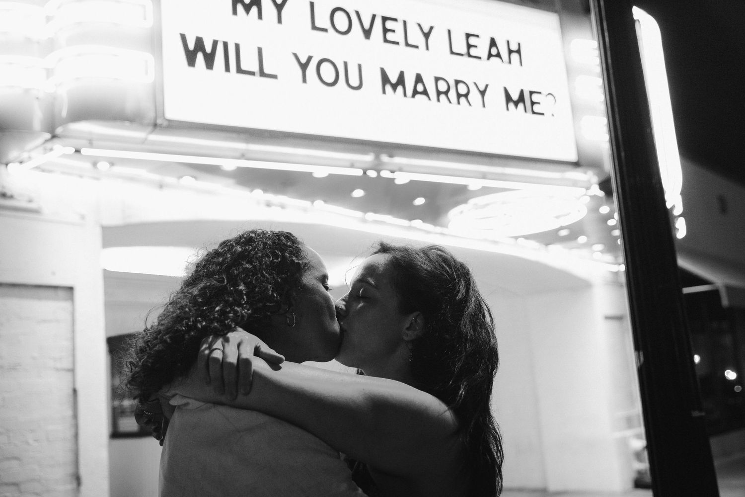 Black and white photo of two women kissing under a theater marriage proposal marquee sign.