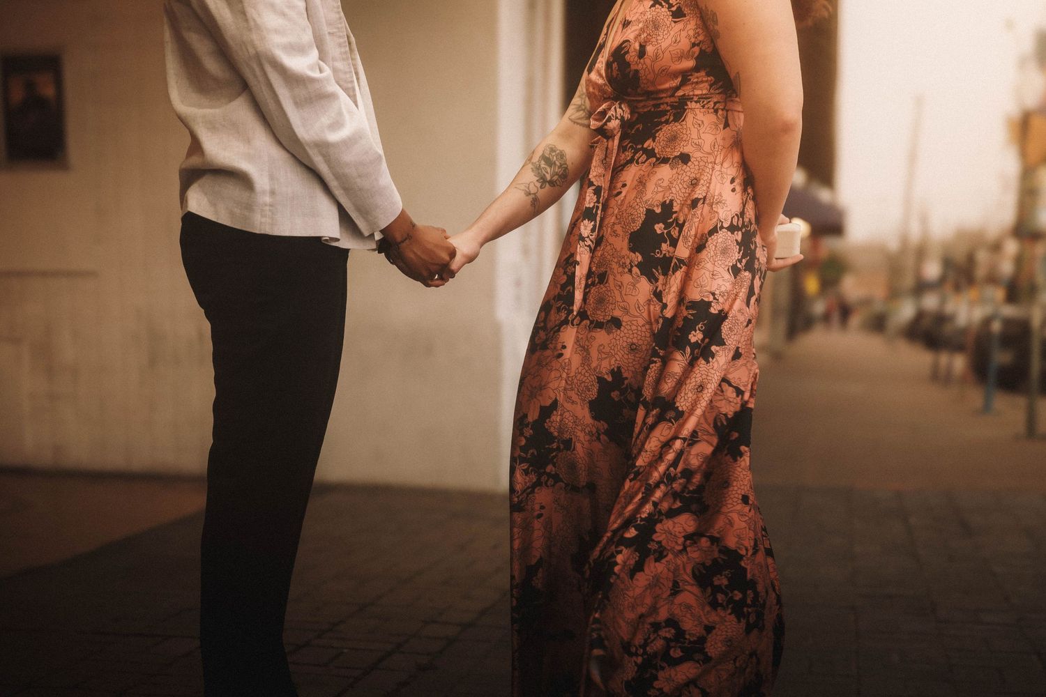 A couple holds hands while walking on a sidewalk, the woman wearing a floral maxi dress with a tattoo visible on her arm.