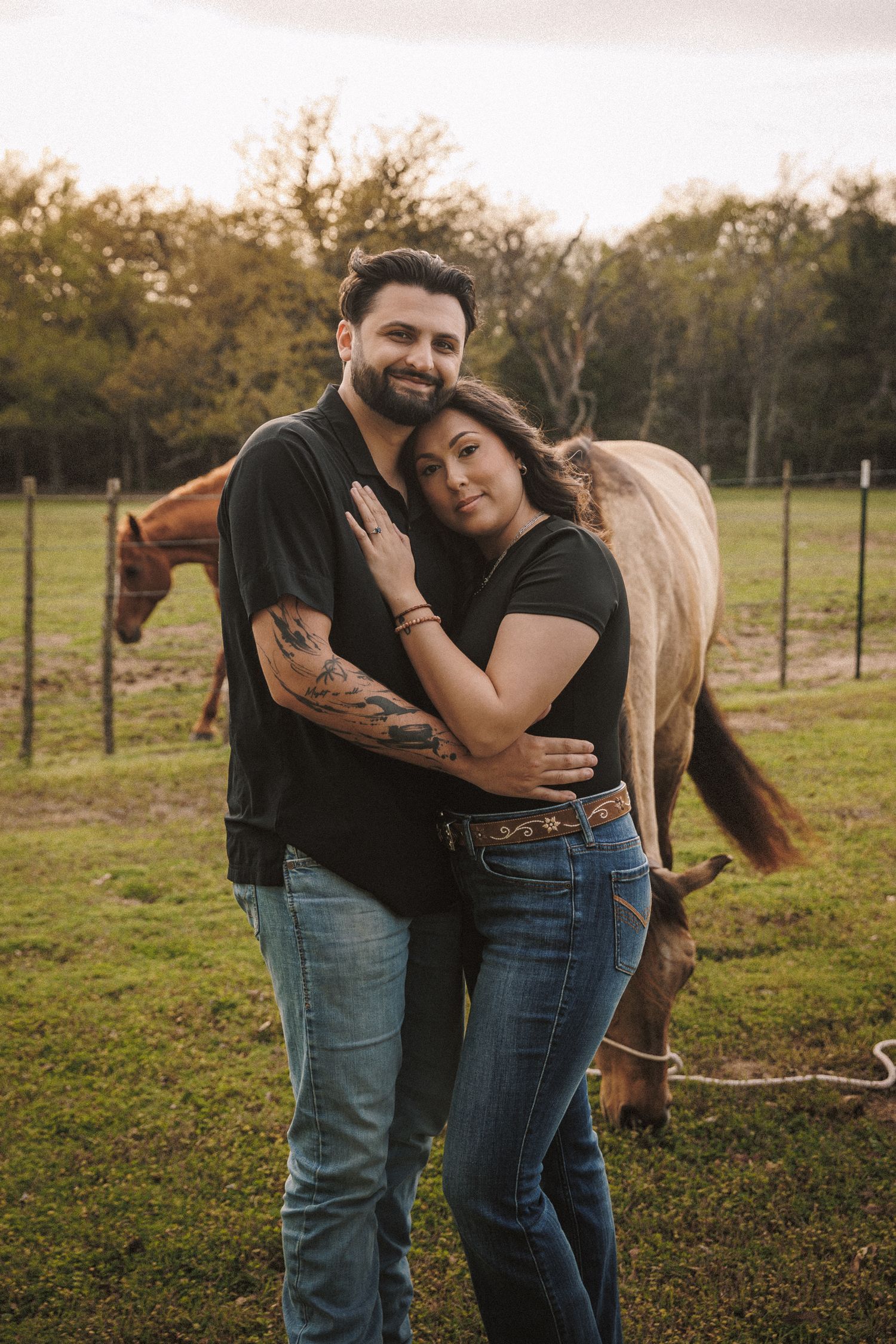 Couple embracing warmly in matching black shirts at sunset on a farm with a horse behind them.