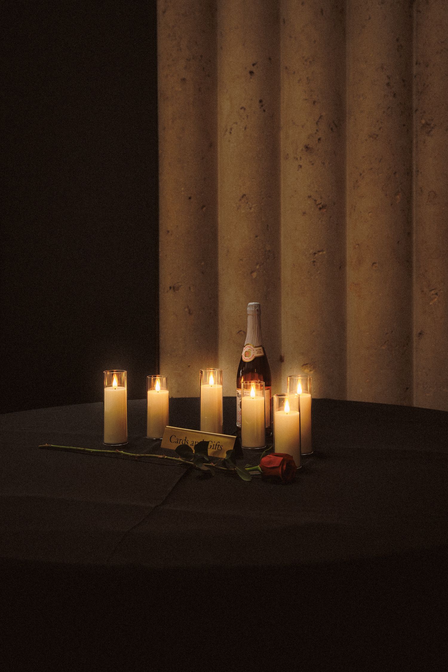 Romantic candlelit display with white candles, a champagne bottle, and a red rose on a dark surface near a column.