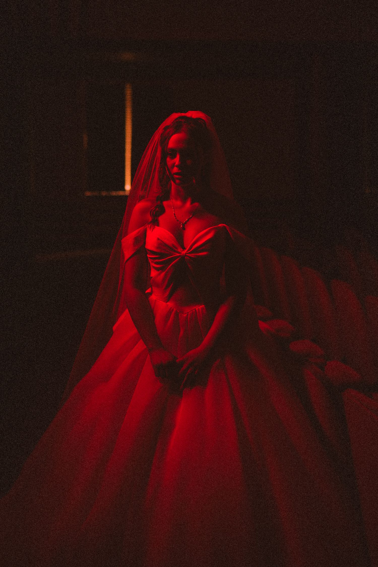 Bride in dramatic red ball gown with veil standing in dark room illuminated by red light.