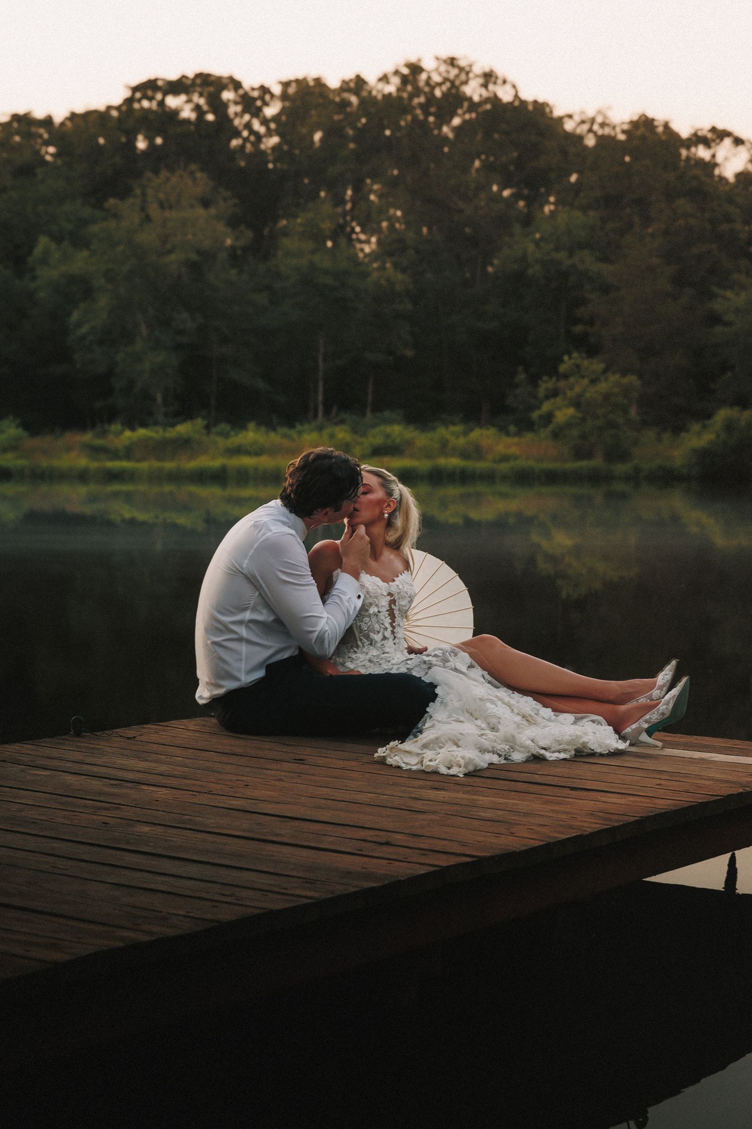 Couple in wedding attire poses romantically on a lakeside dock at dusk.