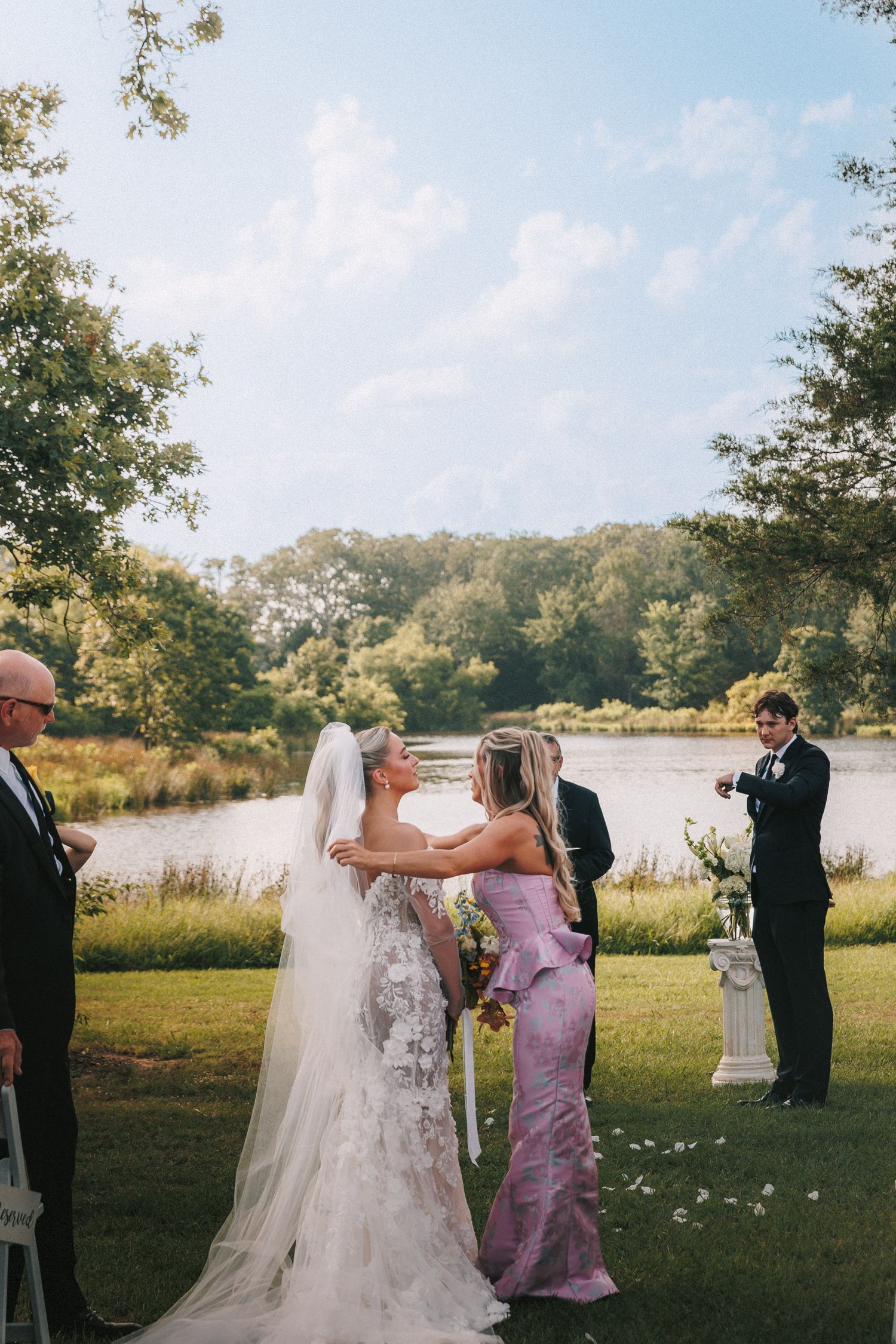 Wedding party members gather on a grassy lawn near a lake during the ceremony.