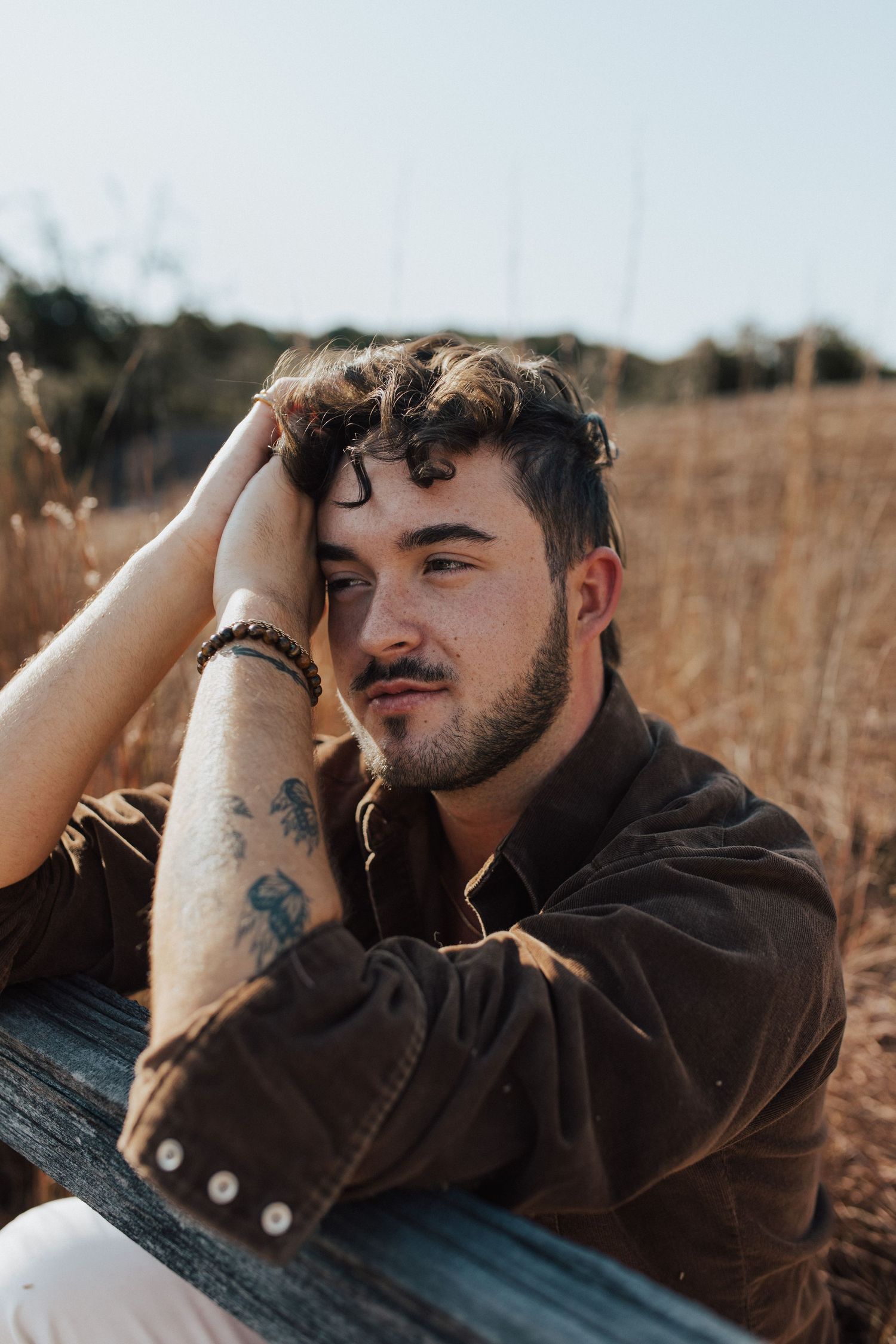 Person wearing brown jacket rests head on wooden surface in golden field during sunset.