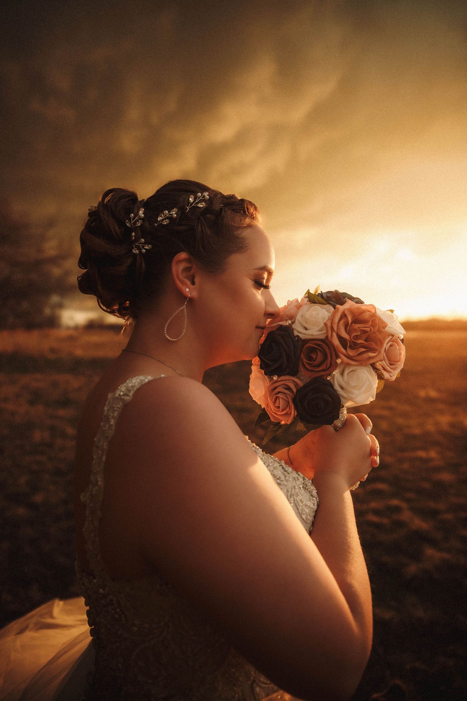 A bride in a wedding dress smells a bouquet of orange and white roses against a warm sunset backdrop.