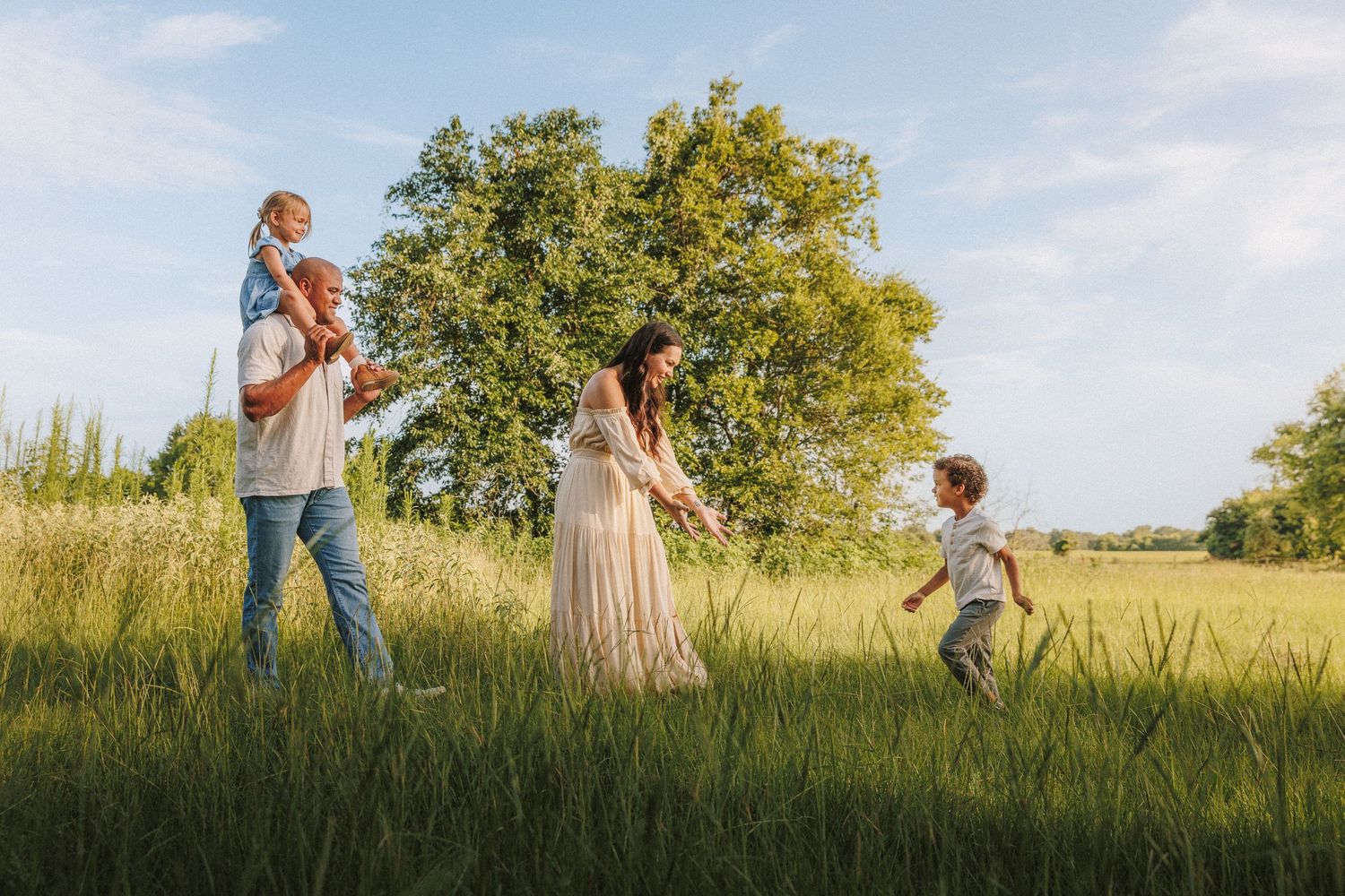 Family running and playing together in a golden sunlit field with tall grass and green trees in the background.