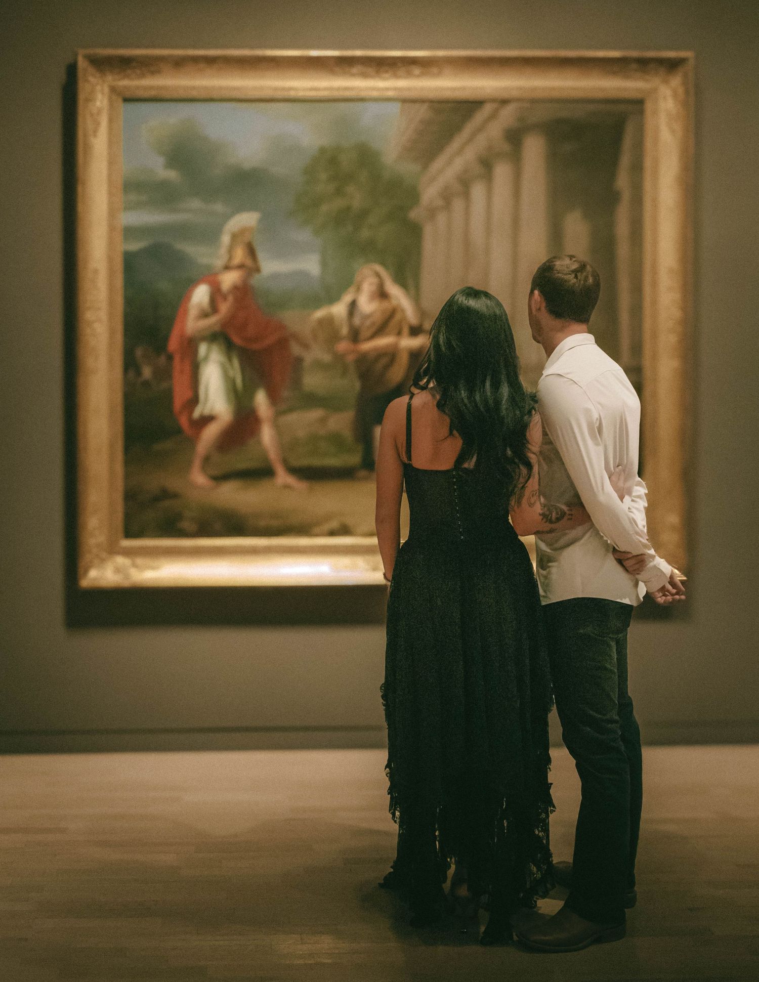 People observe classical paintings in an art gallery exhibit with warm lighting and golden frames on display.