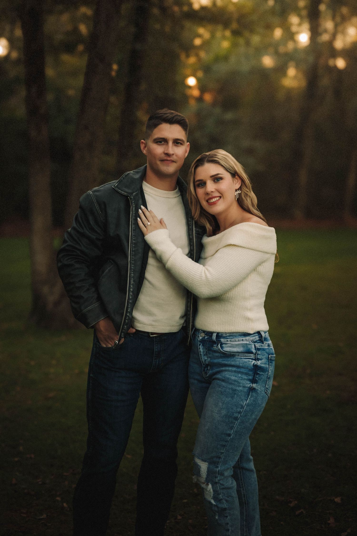 A romantic couple in casual attire share an intimate moment against a wooded sunset backdrop.