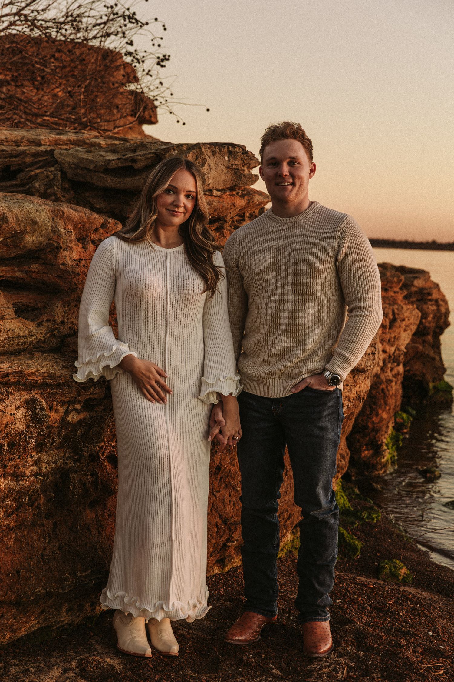 Couple in matching sweaters posing on rocky cliff by water at sunset.