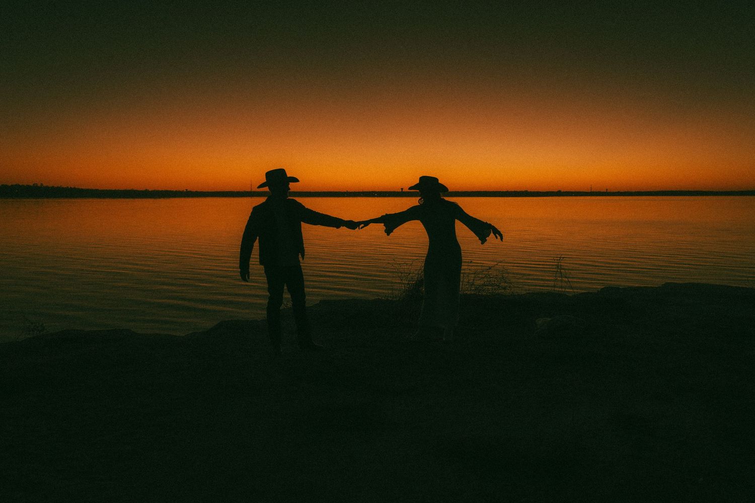 Dark silhouettes of figures against an orange sunset sky reflected on calm water surface.