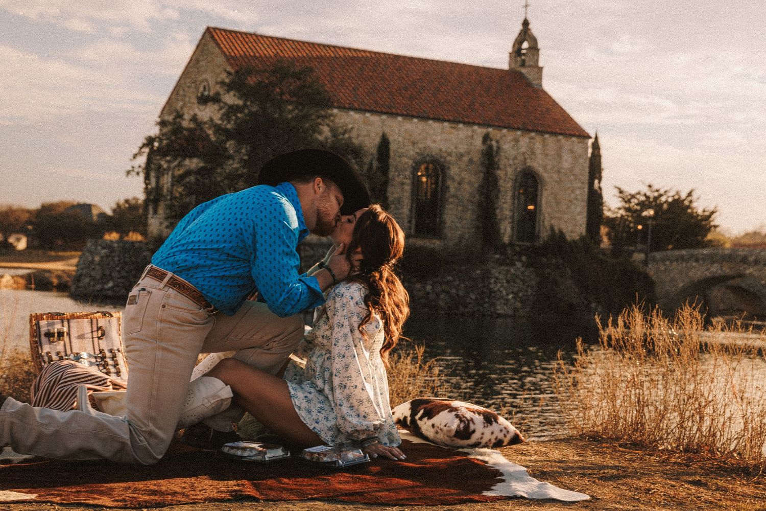 Romantic couple embraces at sunset in front of rustic stone church with warm golden lighting.