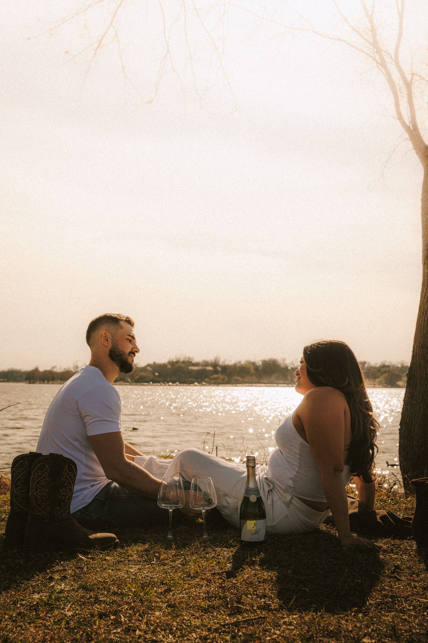 A couple sits together on grass watching a sunset over water.