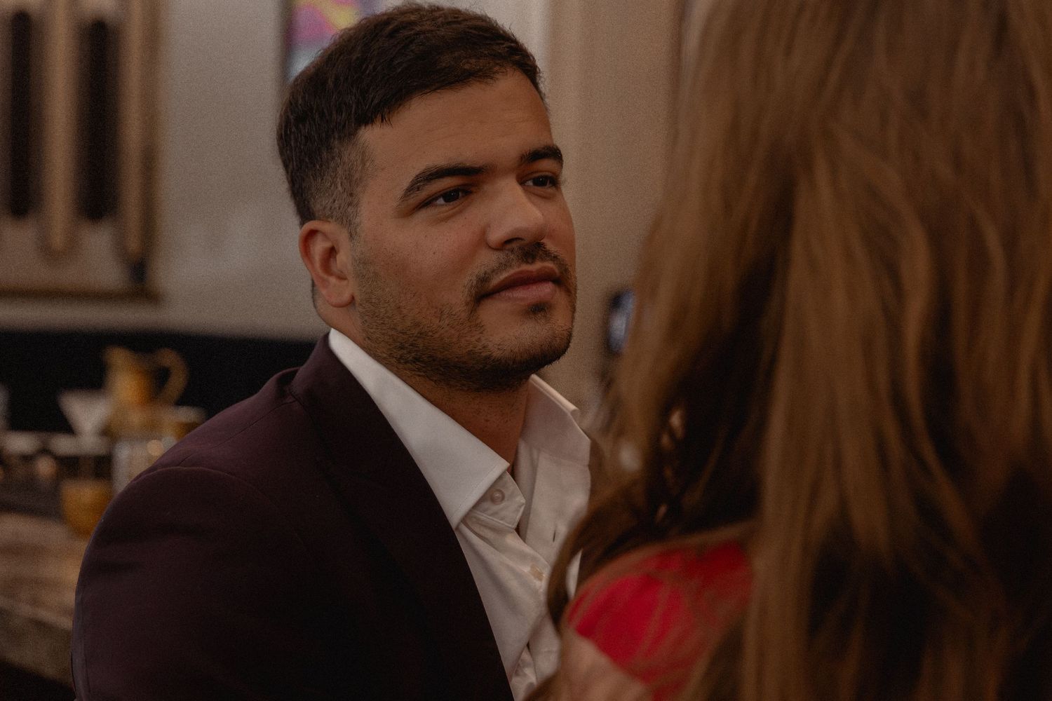 Man in a dark suit gazing romantically at a woman in a red dress indoors.