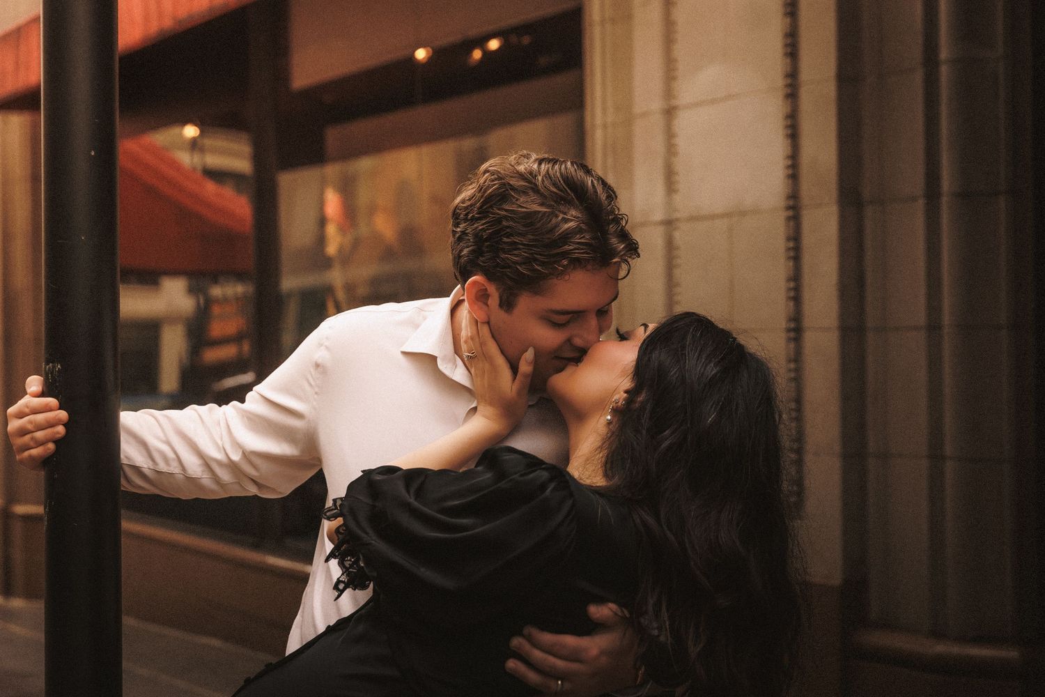A couple shares a romantic kiss outside a warmly lit urban storefront during an engagement photo session.