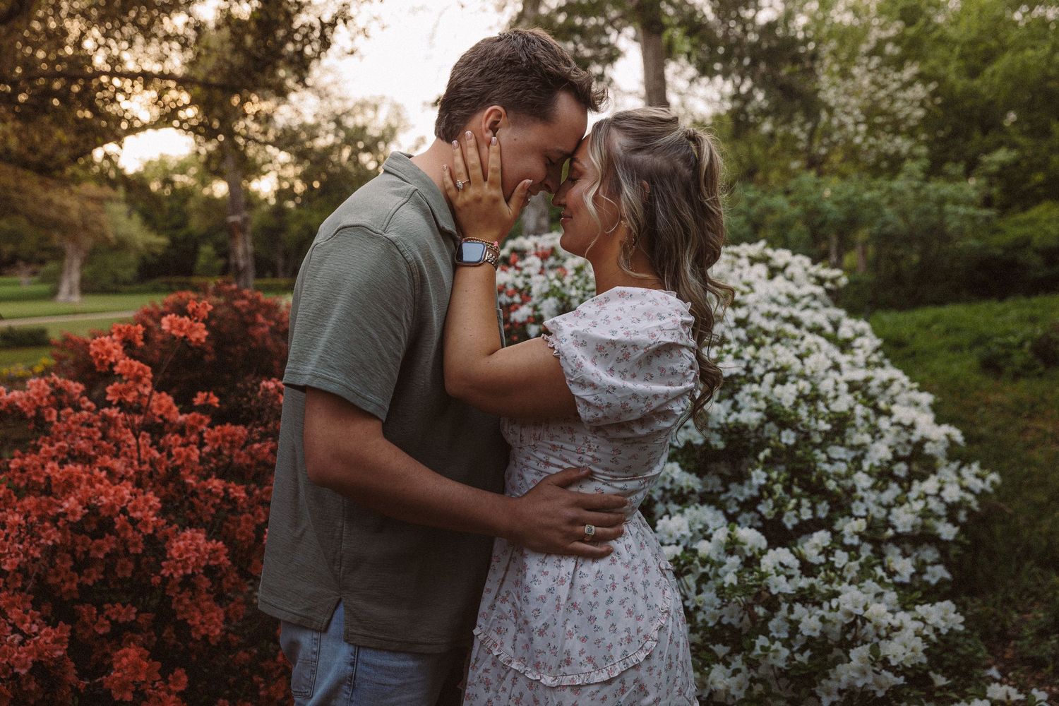 Couple sharing a romantic moment among red and white flowering bushes in a lush garden at golden hour.