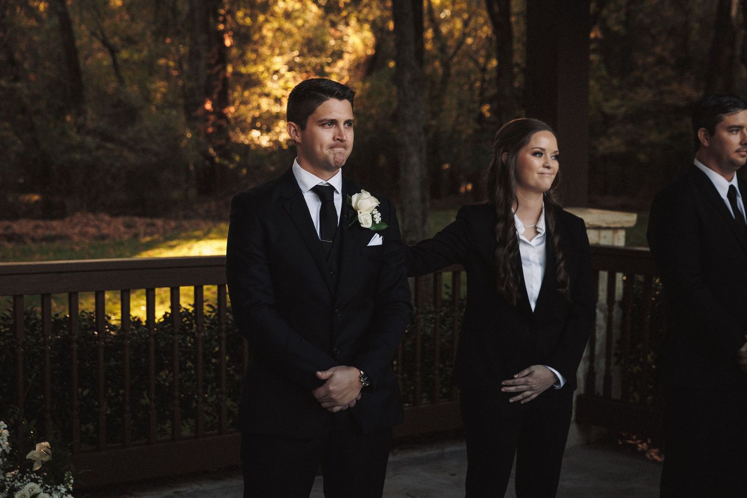 Groom in black tuxedo with white boutonniere stands at altar during outdoor wedding ceremony.