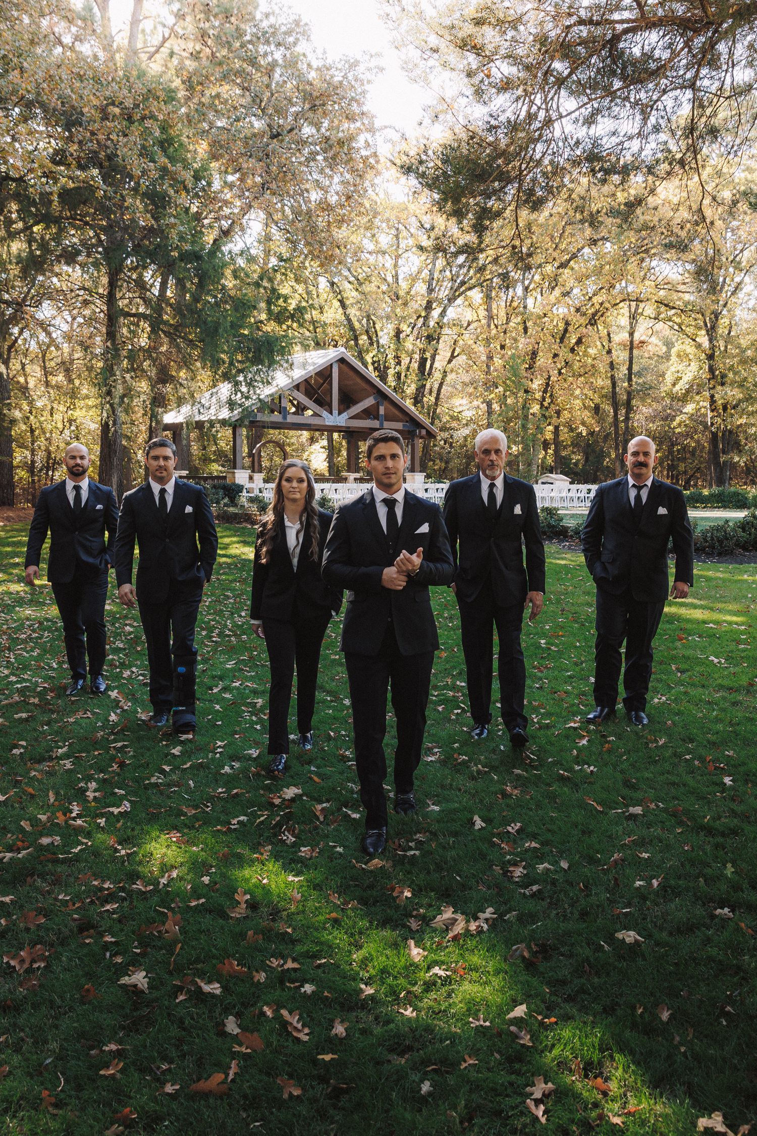 Groomsmen in black suits walk confidently across green lawn with autumn trees and wedding pavilion backdrop.