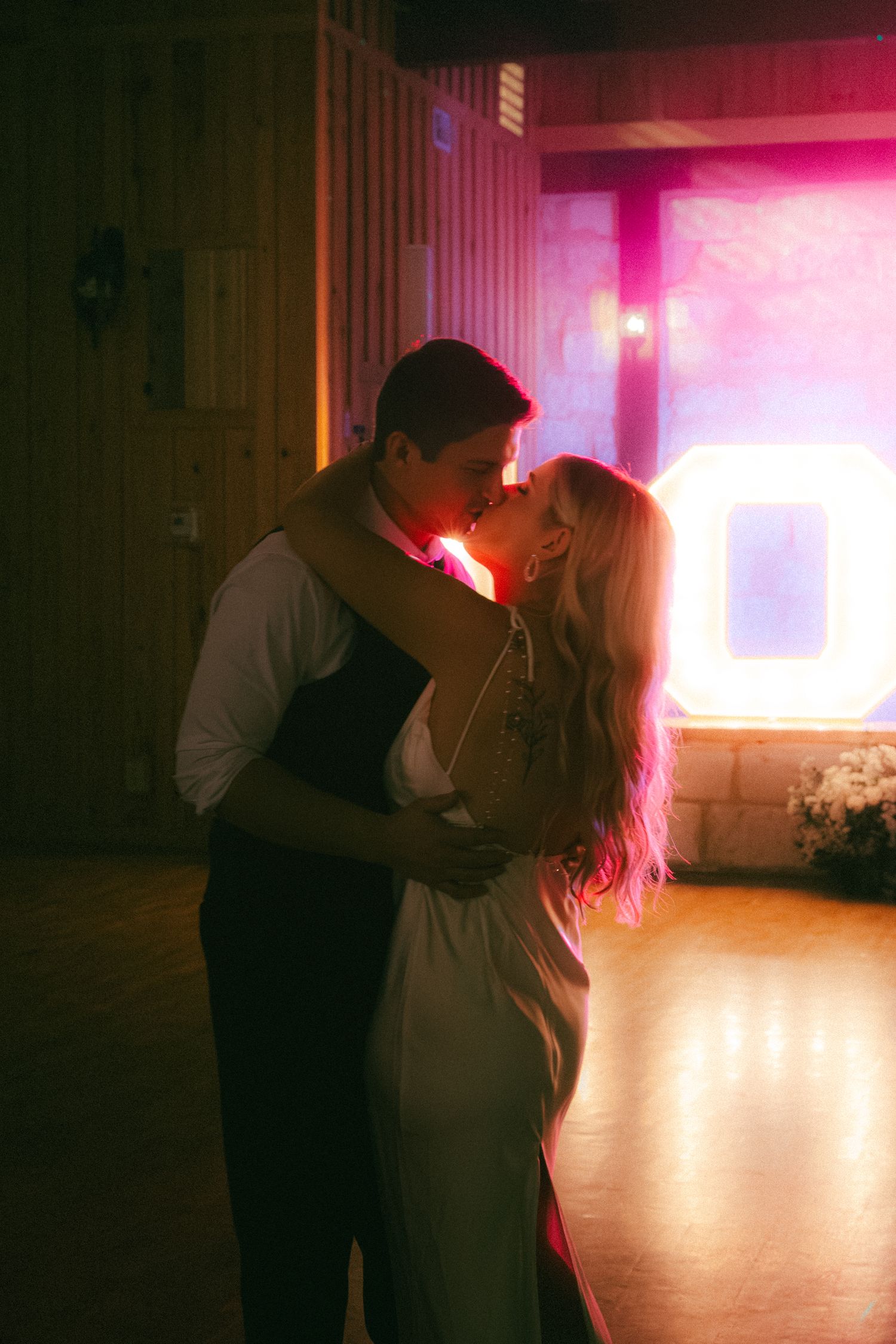 Bride and groom sharing romantic first dance kiss under pink neon lights at wedding.
