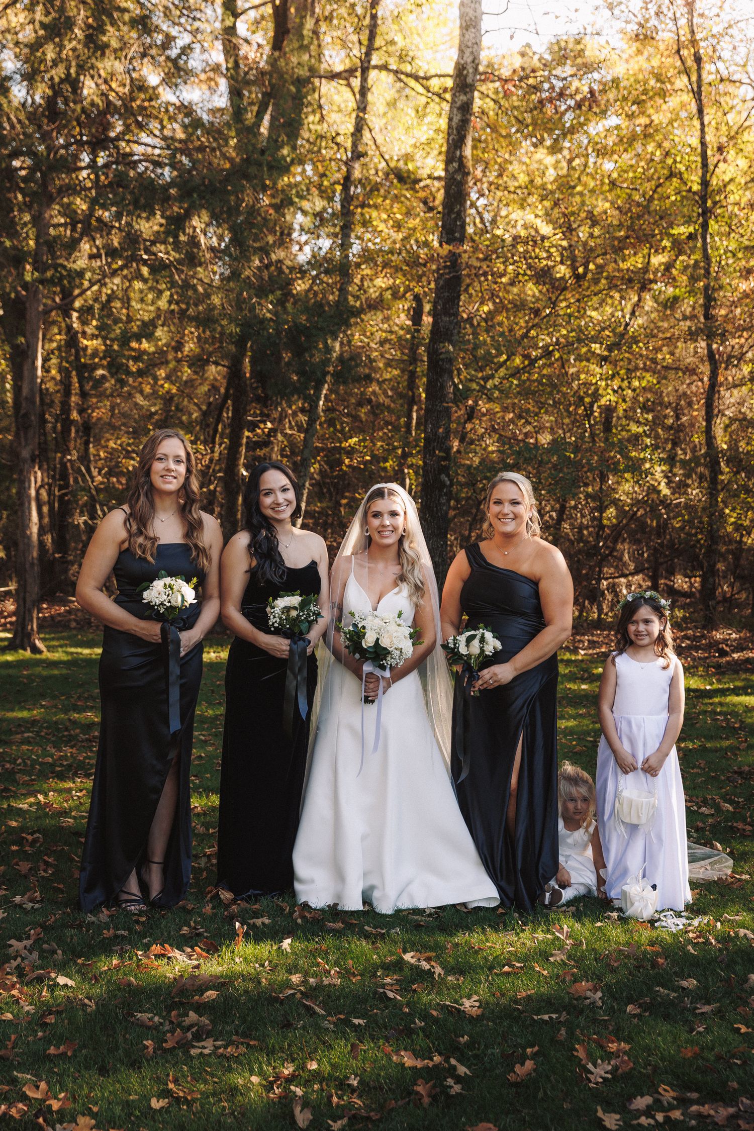 Bride in white gown poses with bridesmaids in black dresses and flower girls in autumn outdoor setting.