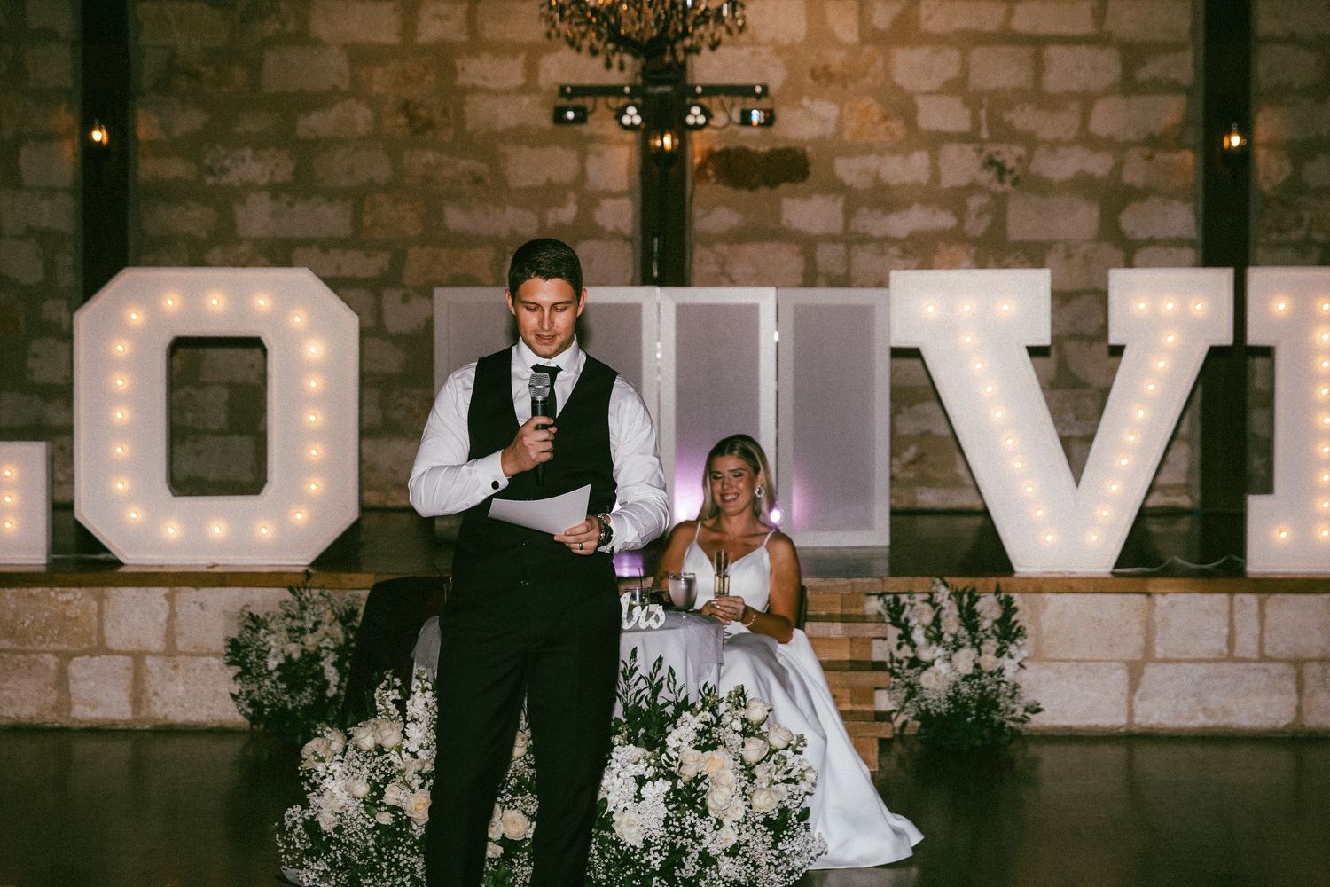 Bride and groom giving speech in front of illuminated LOVE marquee letters at wedding reception.