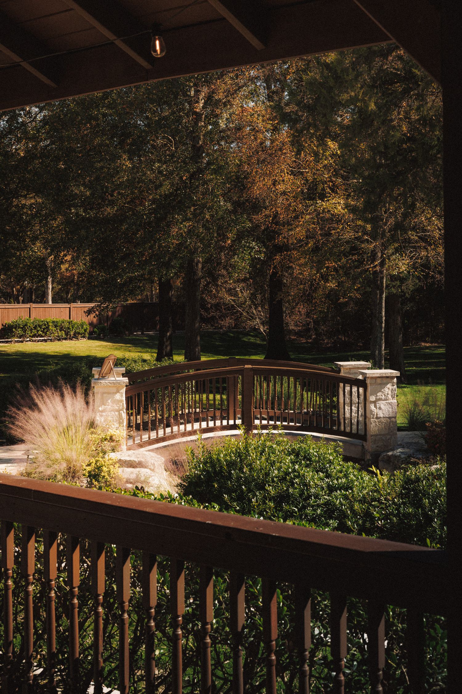 Elegant wooden bridge with stone pillars surrounded by lush green shrubs and autumn trees in a serene garden setting.