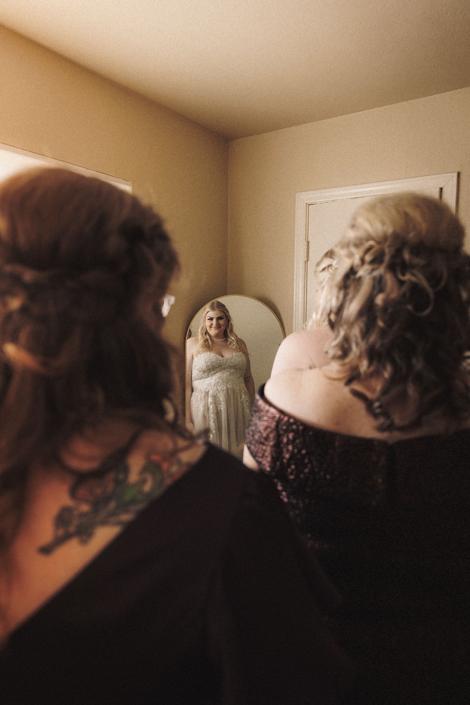 Bride admiring her wedding dress reflection in mirror while bridesmaid with tattoos assists her.