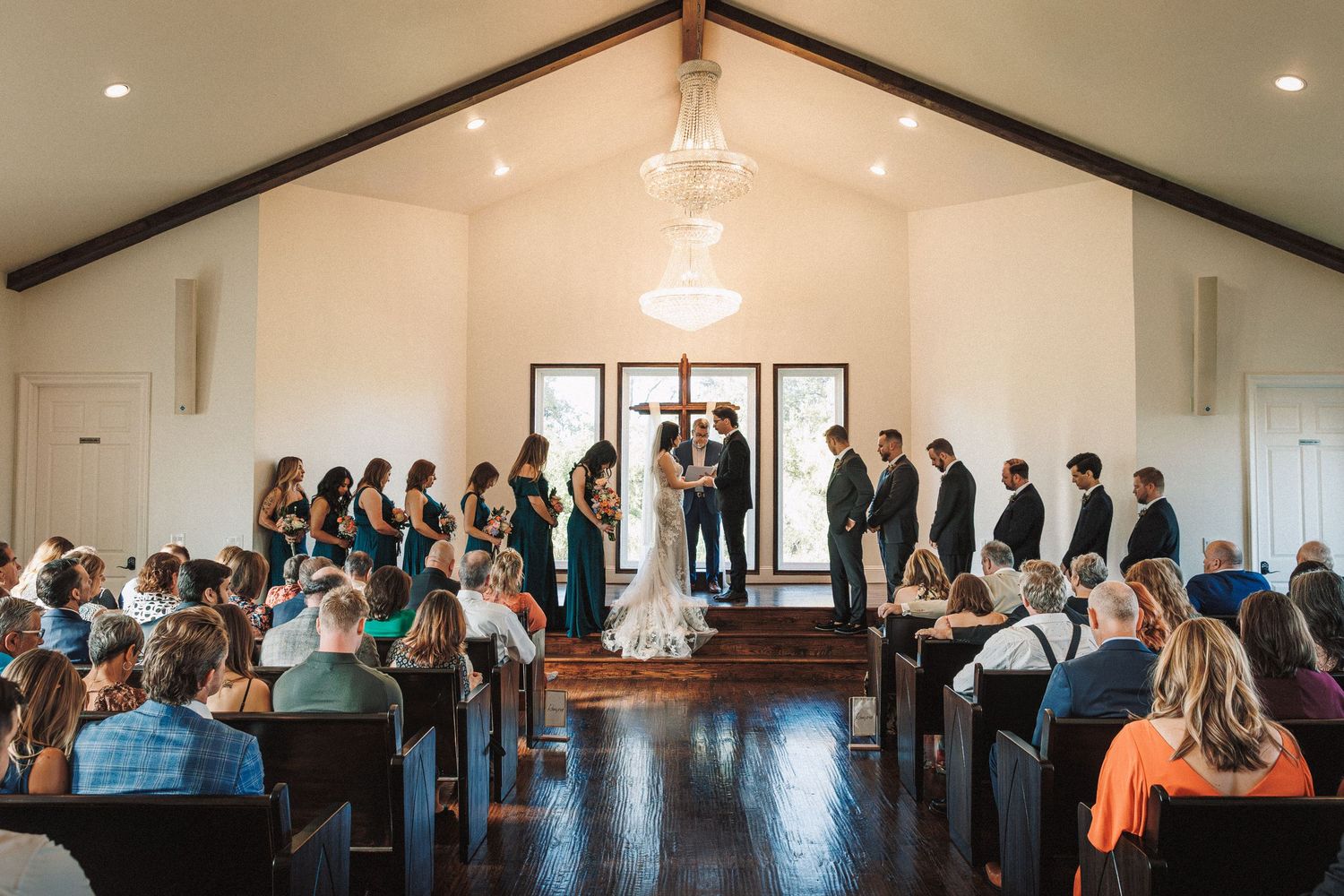 Wide shot of a wedding ceremony inside a white chapel with chandelier, bridal party, and seated guests.