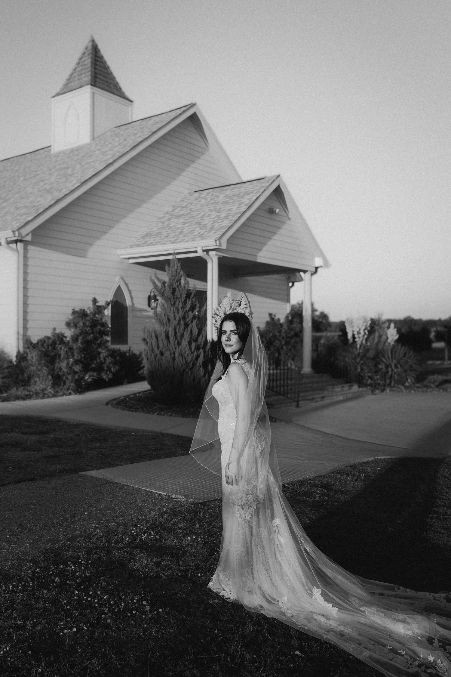Black and white photo of a bride in a lace gown and veil posing gracefully outside a white chapel at dusk.