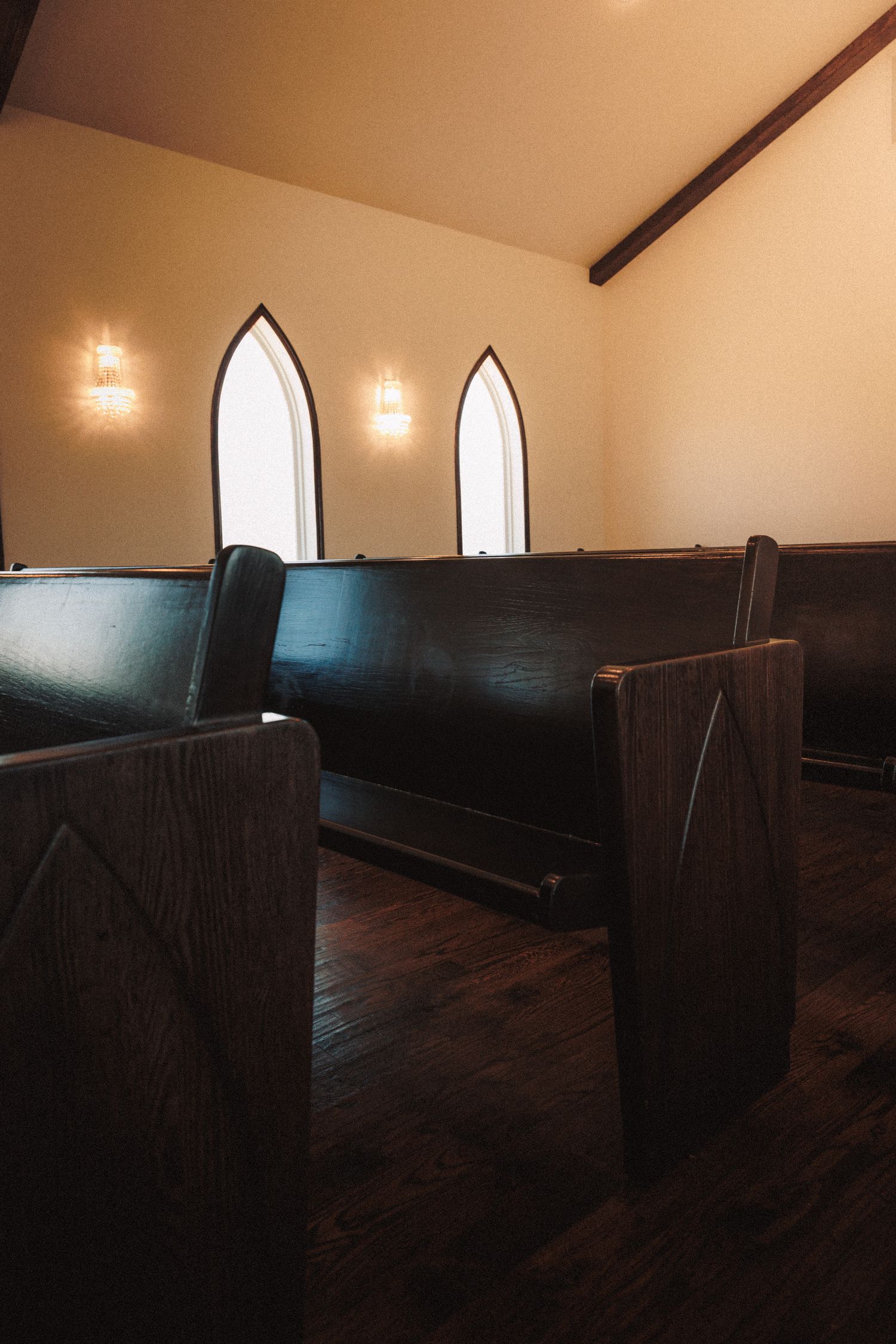 Rustic church interior with dark wooden pews, hardwood floors, and gothic arched windows with warm wall sconces.
