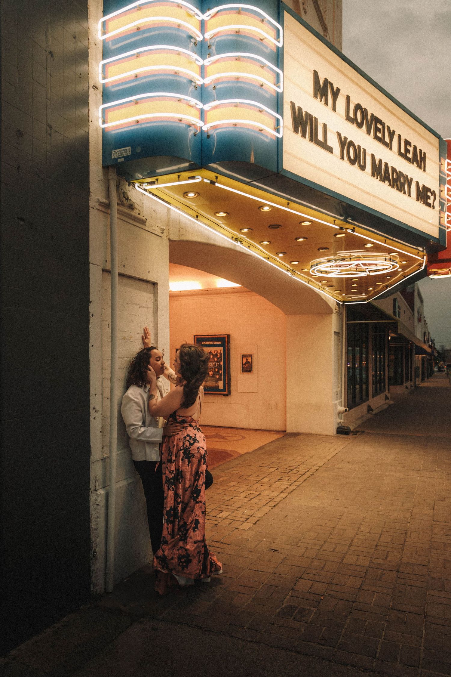 Man and woman embrace against theater wall under neon marriage proposal marquee sign at dusk.