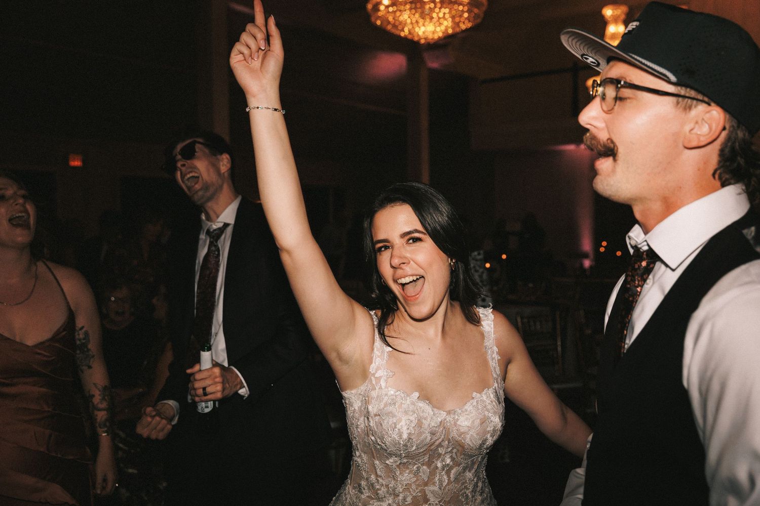 Bride in lace dress dancing joyfully with guests at wedding reception under warm lights.