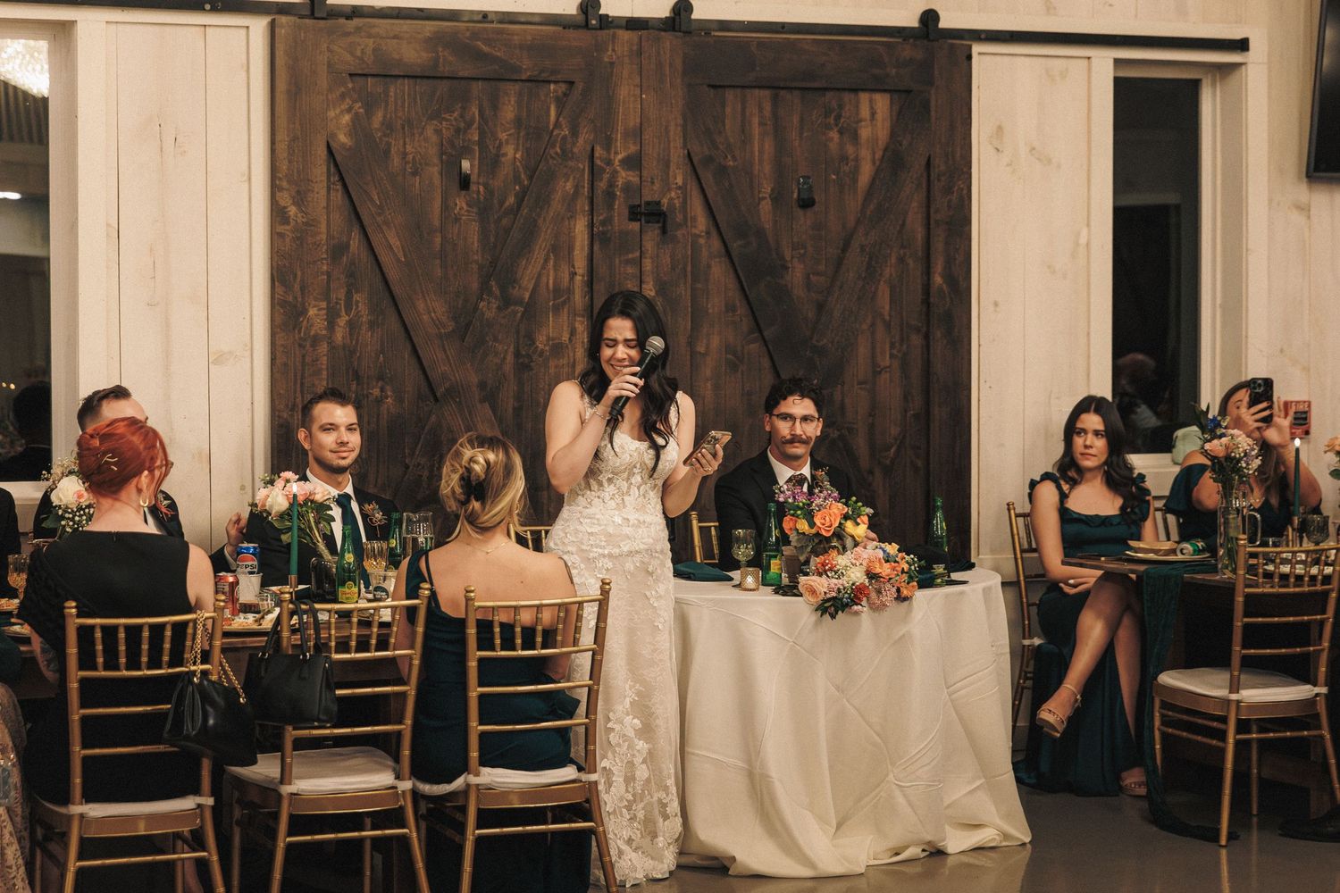 Bride in lace gown speaks at sweetheart table during wedding reception in rustic barn venue with barn doors.