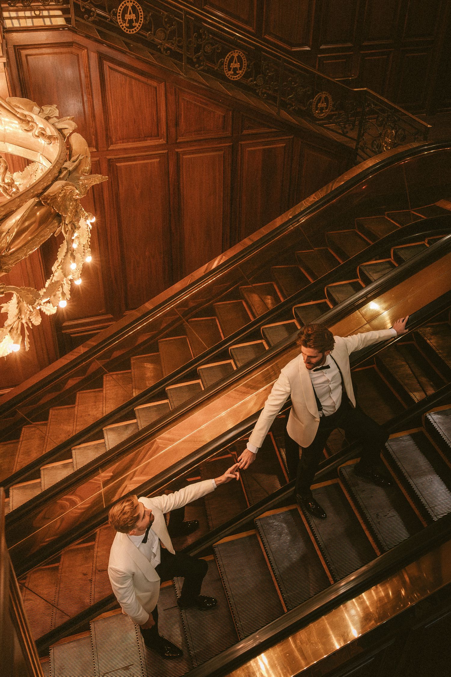 Aerial view of two grooms in white tuxedos holding hands on ornate wooden escalator.