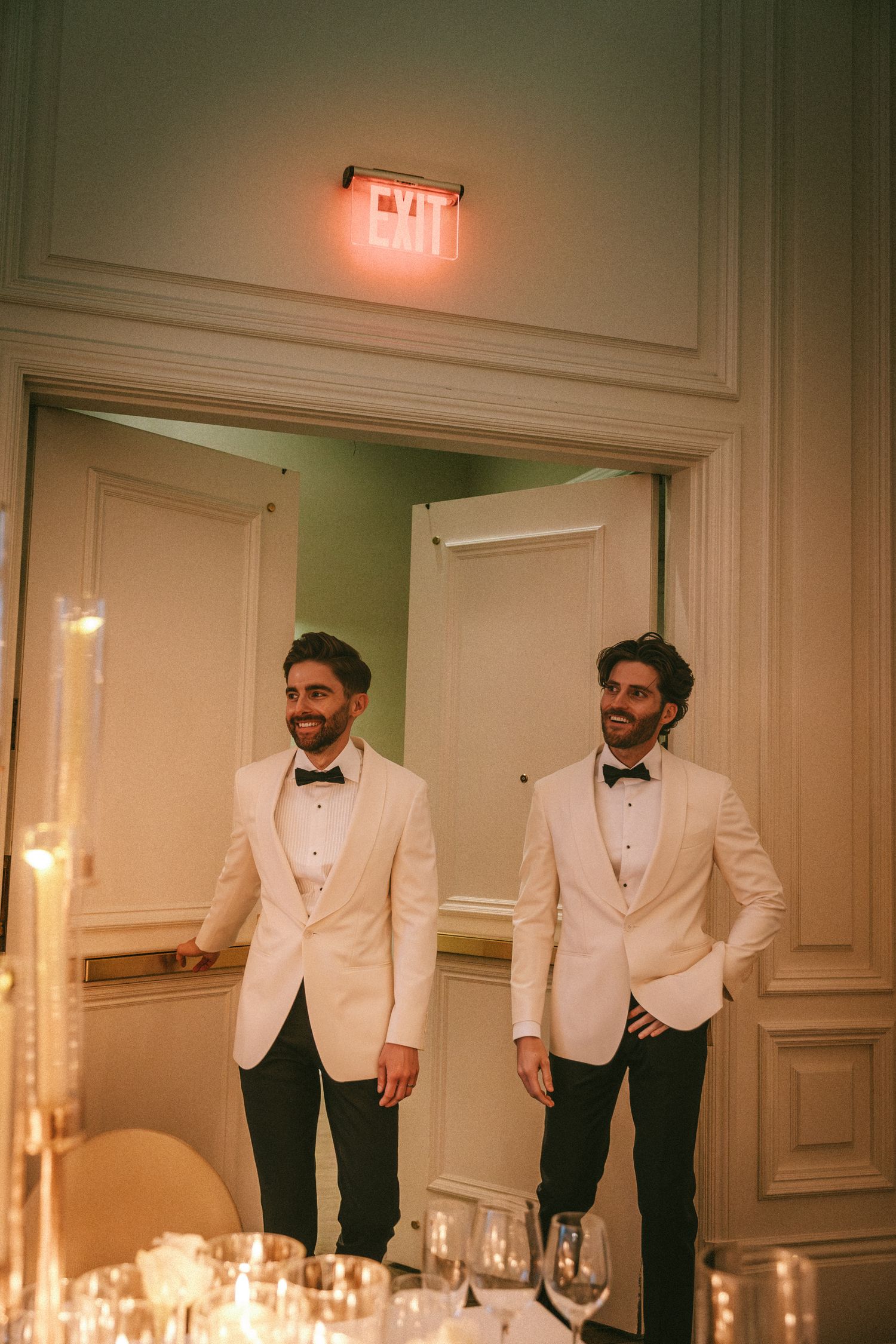 Two grooms in white tuxedo jackets and bow ties smiling in elegant wedding reception venue doorway.