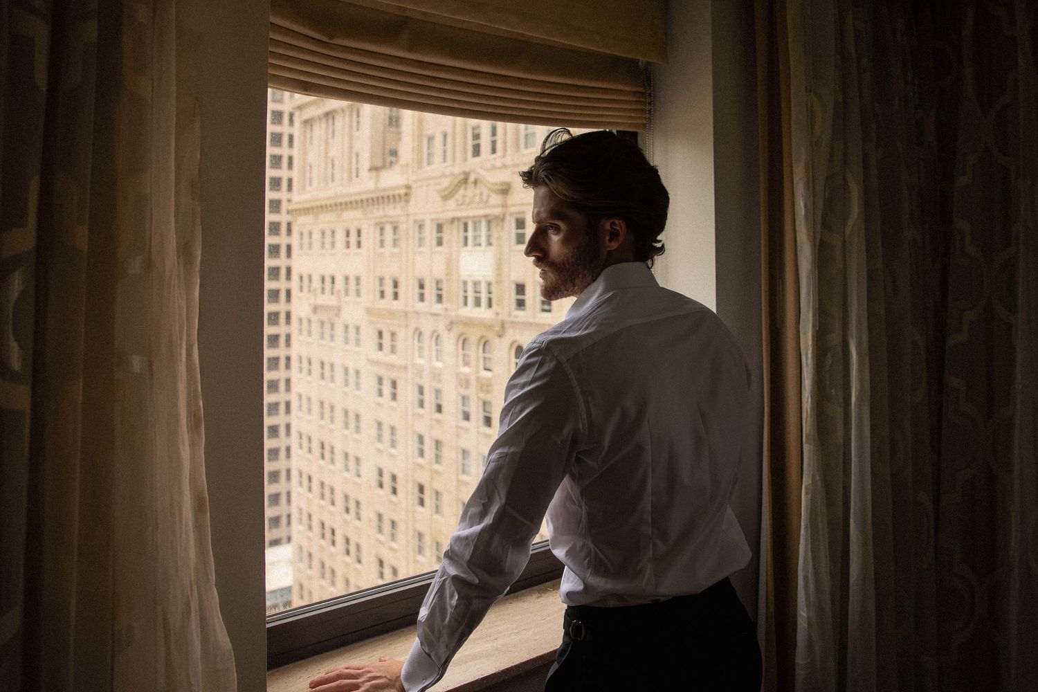 Groom in white dress shirt gazing out hotel window at city skyline before wedding.