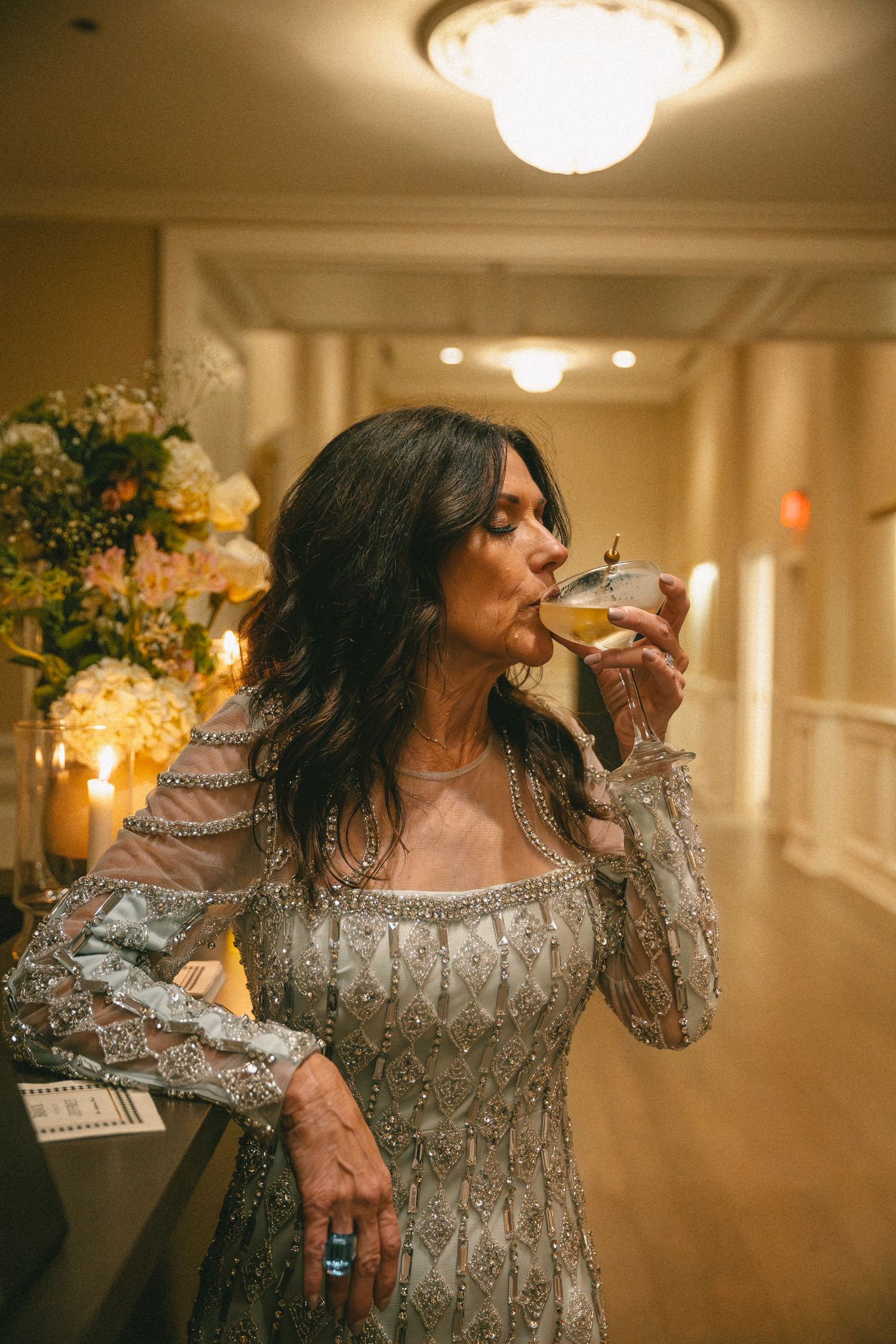 An elegantly dressed woman in a silver beaded gown sips a cocktail in a warmly lit hallway.