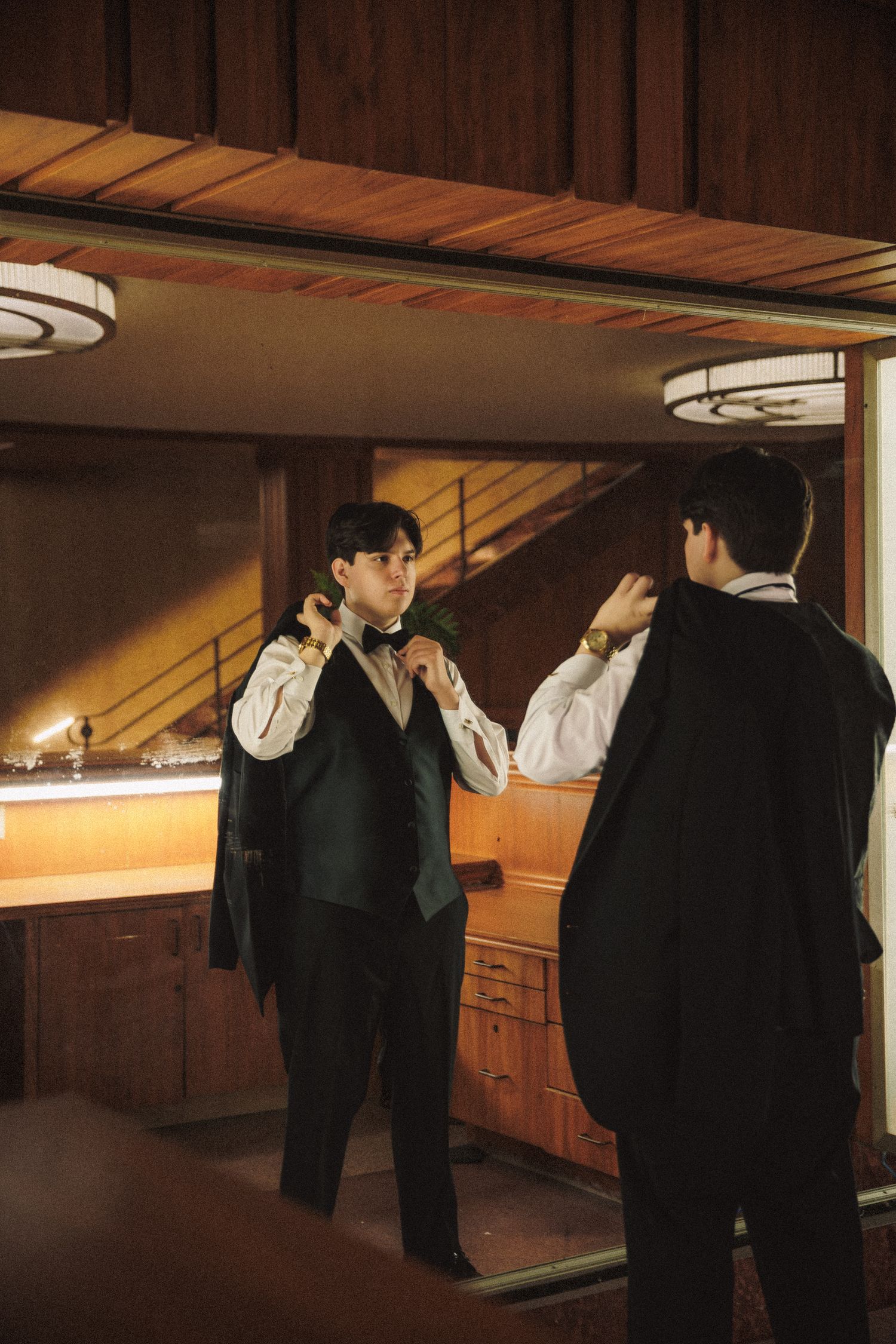 Groom adjusting bow tie in mirror reflection inside warmly lit wood-paneled room.