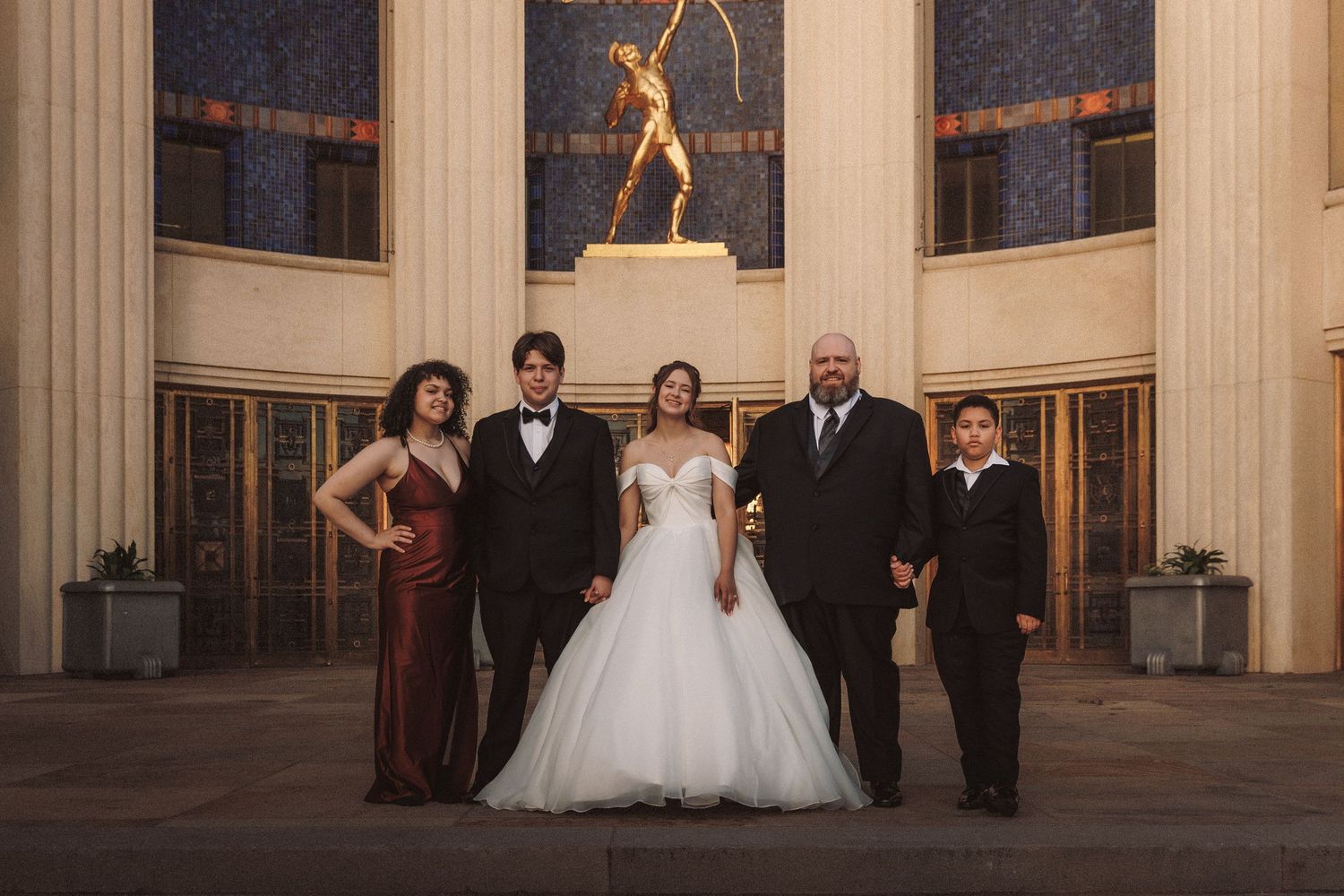 Wedding party posing before golden statue in grand art deco venue with bride in white ballgown.