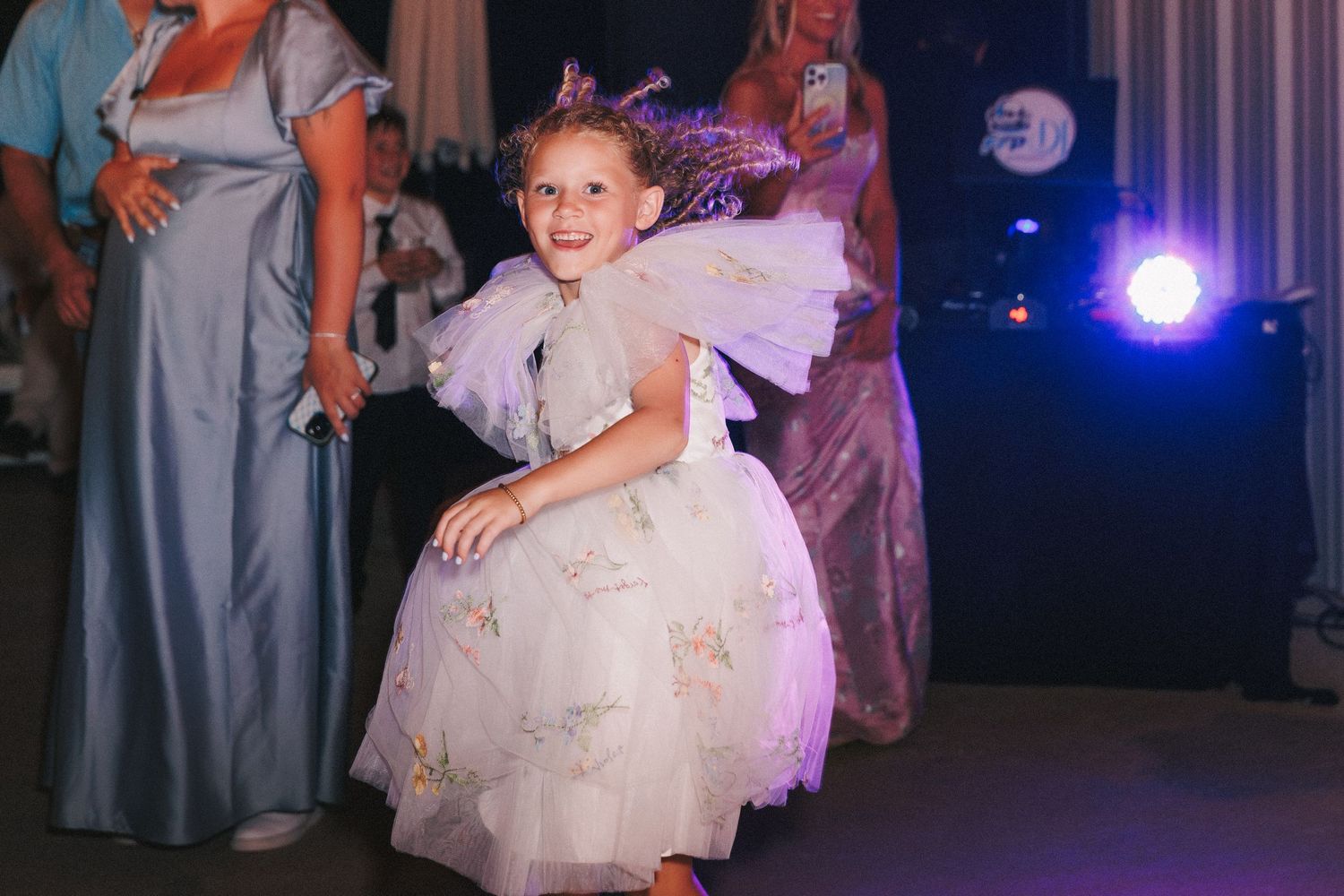 A little girl in a white tulle dress twirls on a dance floor with purple lighting.