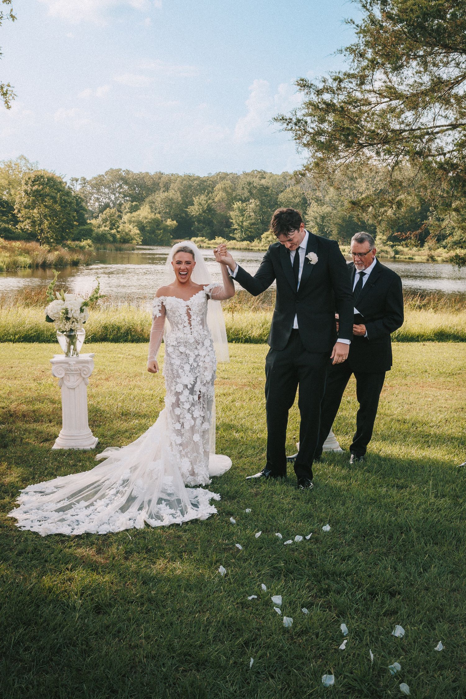 Wedding party walking and celebrating on grassy field with long lace train trailing behind.