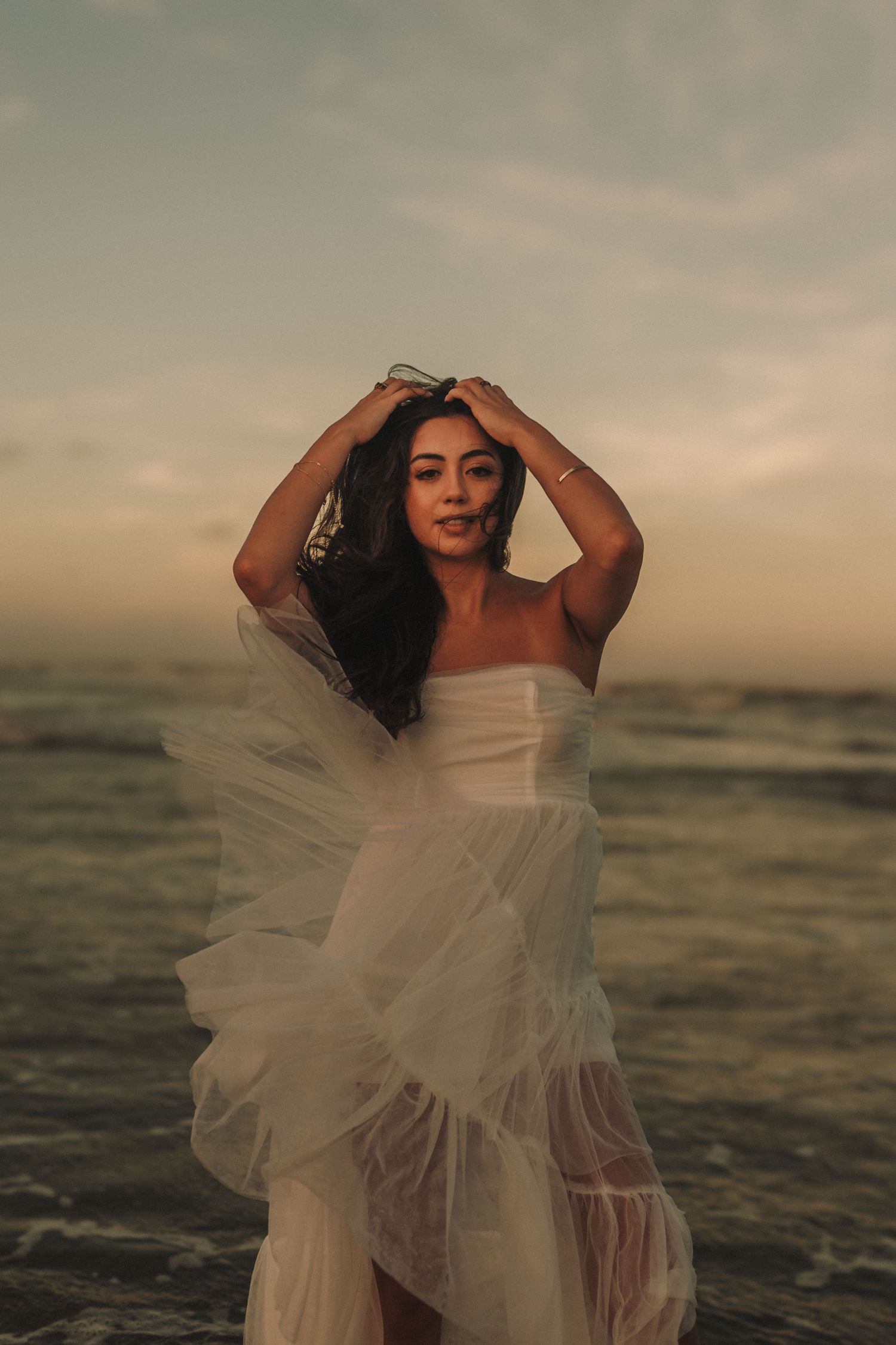 A graceful figure in a flowing white dress poses against dramatic ocean waves at sunset, creating an ethereal scene.